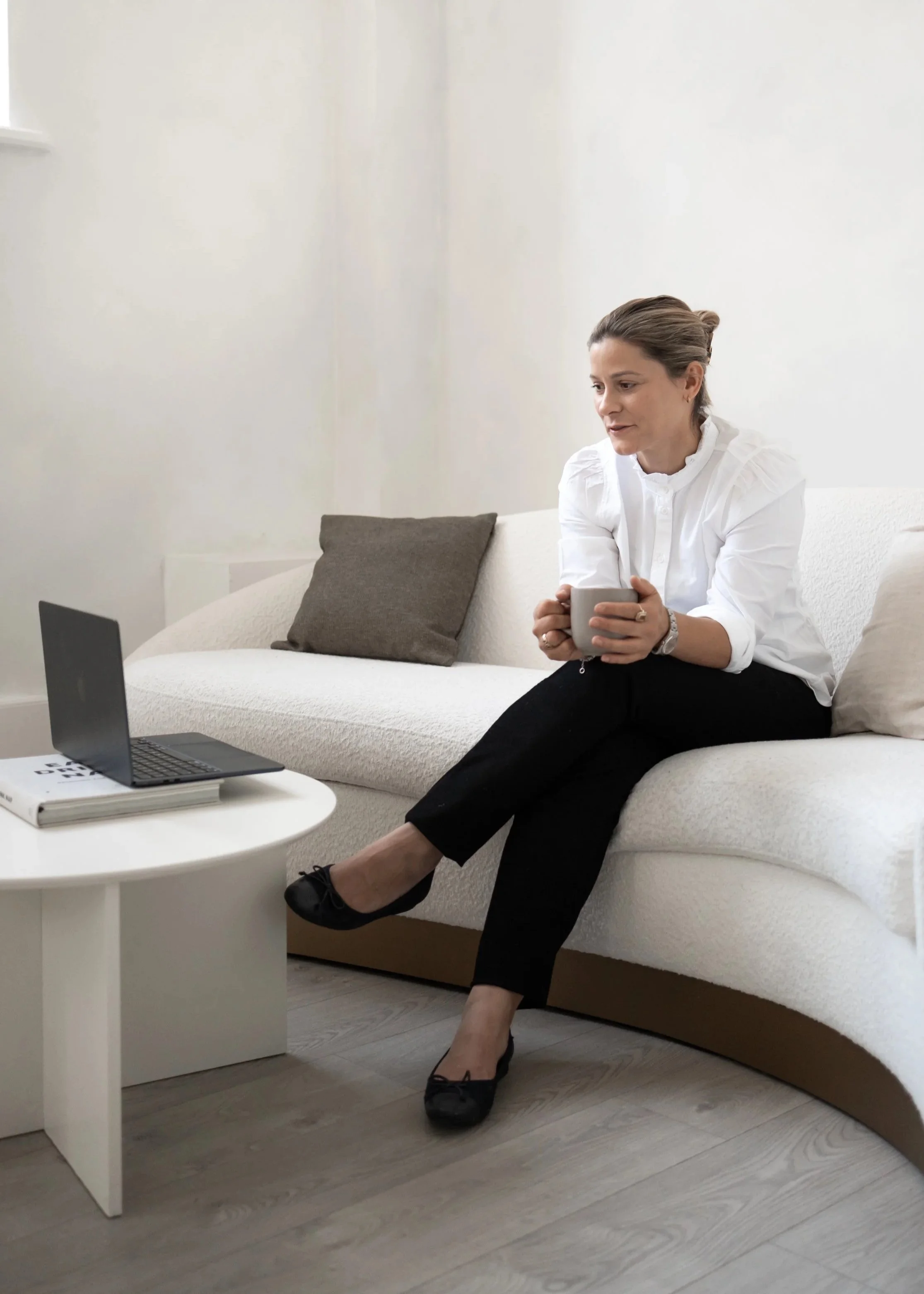 Health Coach and Reiki Practitioner Plum sitting on a white couch holding a coffee mug, with a laptop on a small white table in front of her. The room has a neutral colour palette with light-coloured wooden floors and a gray pillow on the couch.