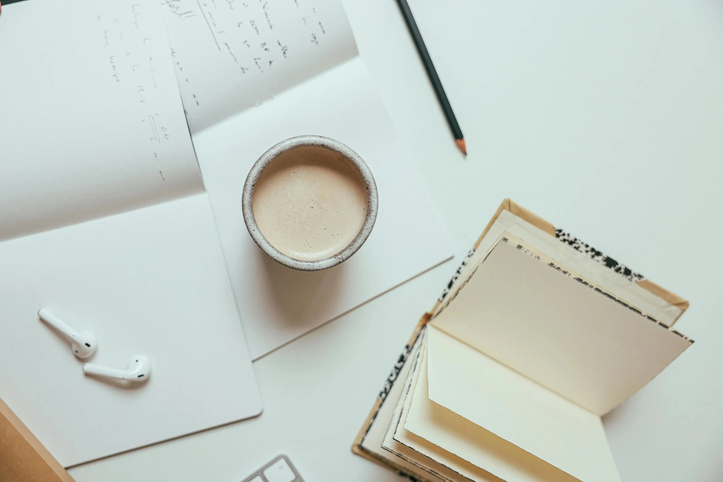 Top-down view of a workspace with a white surface, featuring a partially visible laptop with white wireless earbuds and charging case, a stack of books or notebooks, a pencil, a piece of handwritten paper, and a beige coffee mug filled with a frothy beverage.