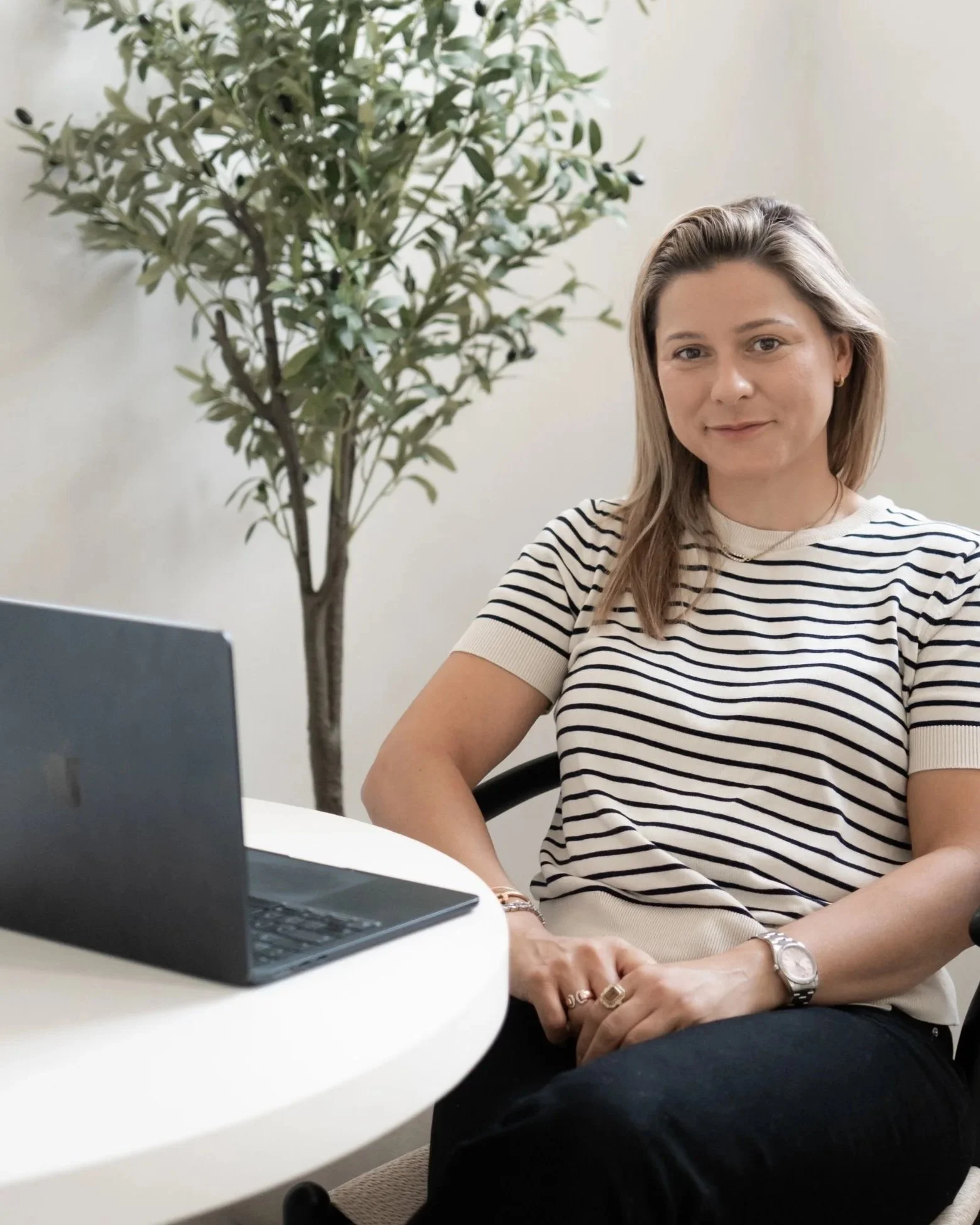 Health Coach and Reiki MasterPlum, wearing a white and black striped shirt, sitting at a white desk with a silver watch on her left wrist, in front of a laptop, in a room with a tall green plant in the background.