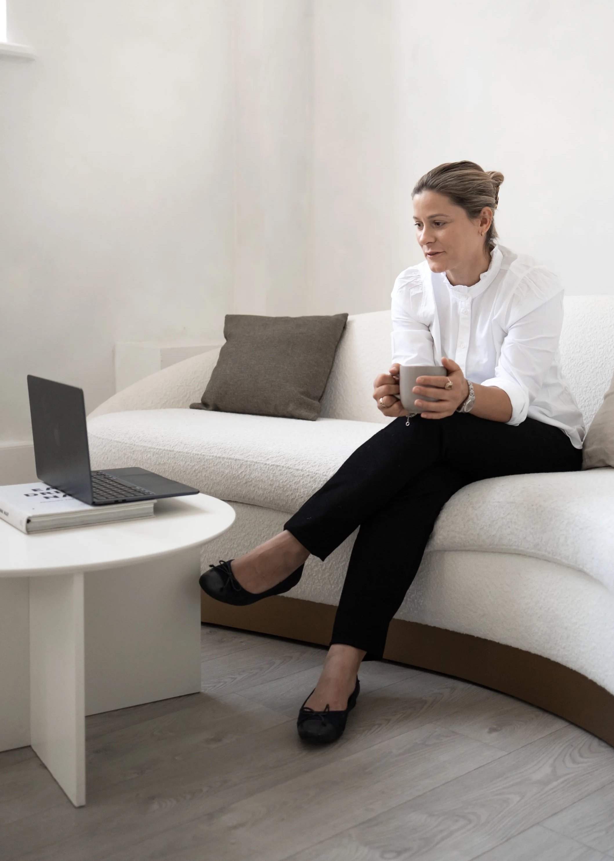 Woman sitting on a white sofa holding a gray mug, working from home with a laptop on a small white table nearby, in a minimalist living room with gray flooring and a gray pillow on the sofa.