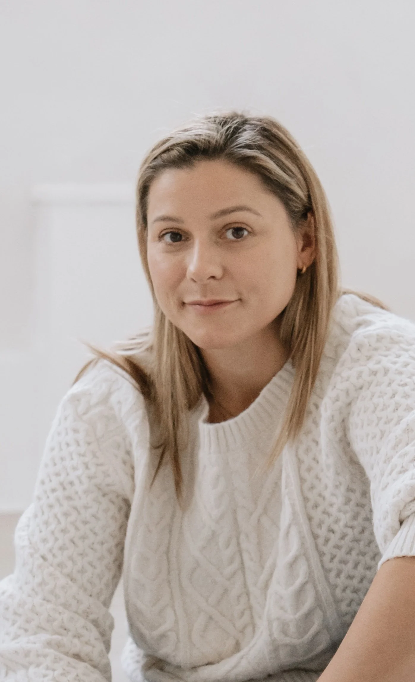 A woman with light brown hair and fair skin wearing a white, textured, knit sweater, sitting indoors against a plain white background.