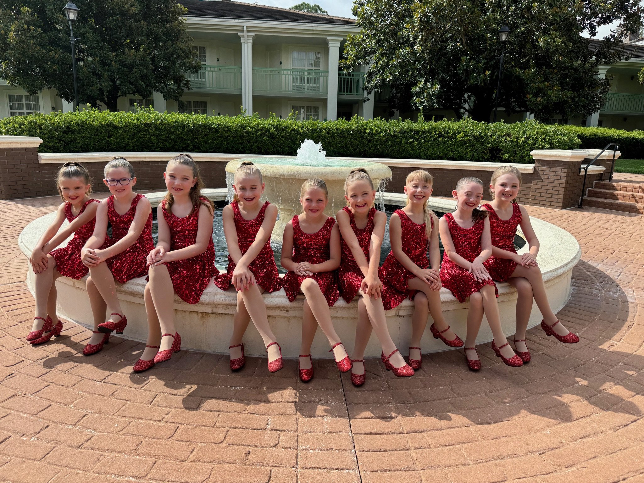 A group of ten young girls wearing red sequin dresses and matching shoes sitting on the edge of a circular fountain in an outdoor courtyard.