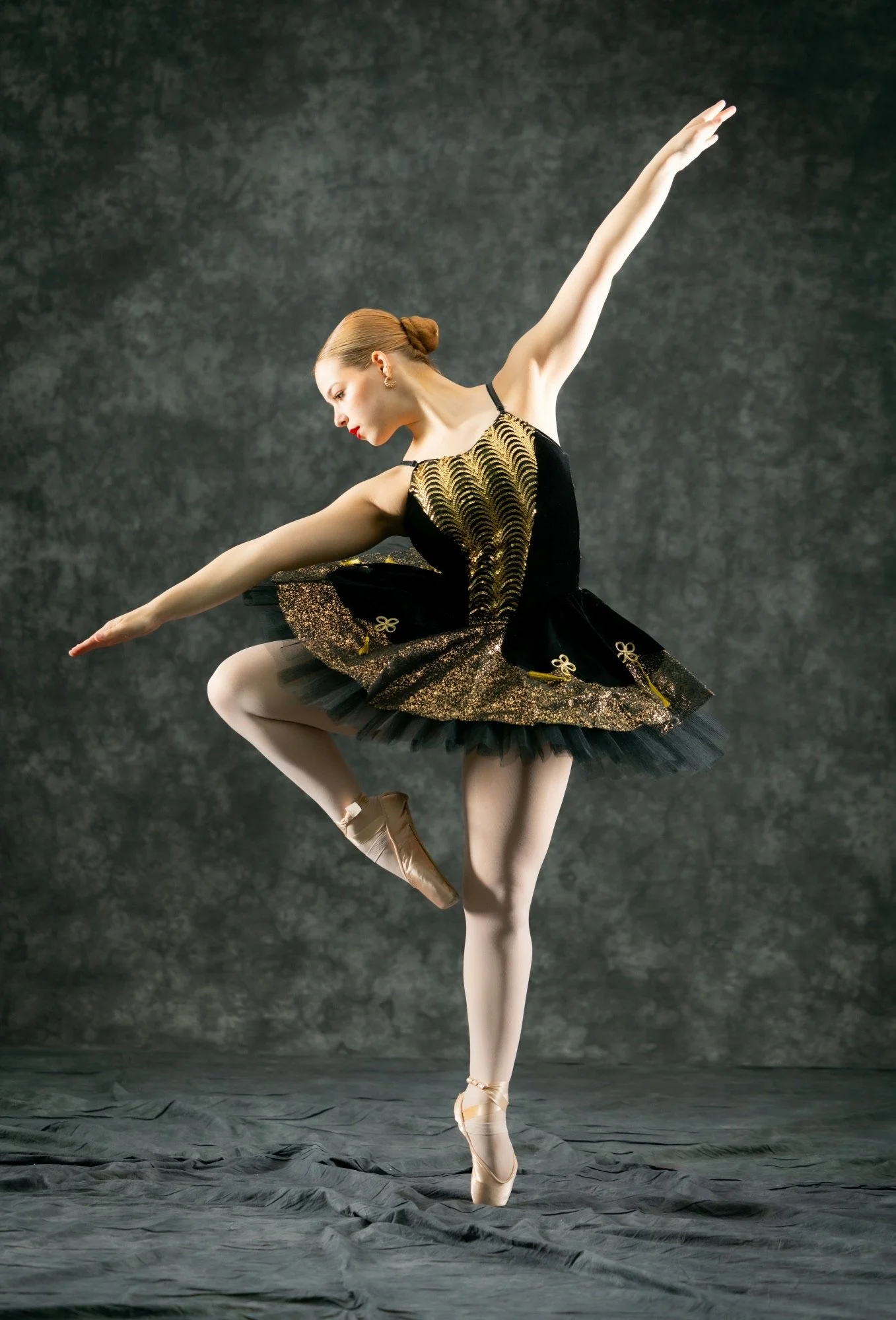 A ballet dancer in a black and gold tutu performs a pose, standing on one leg with the other lifted and arms extended.