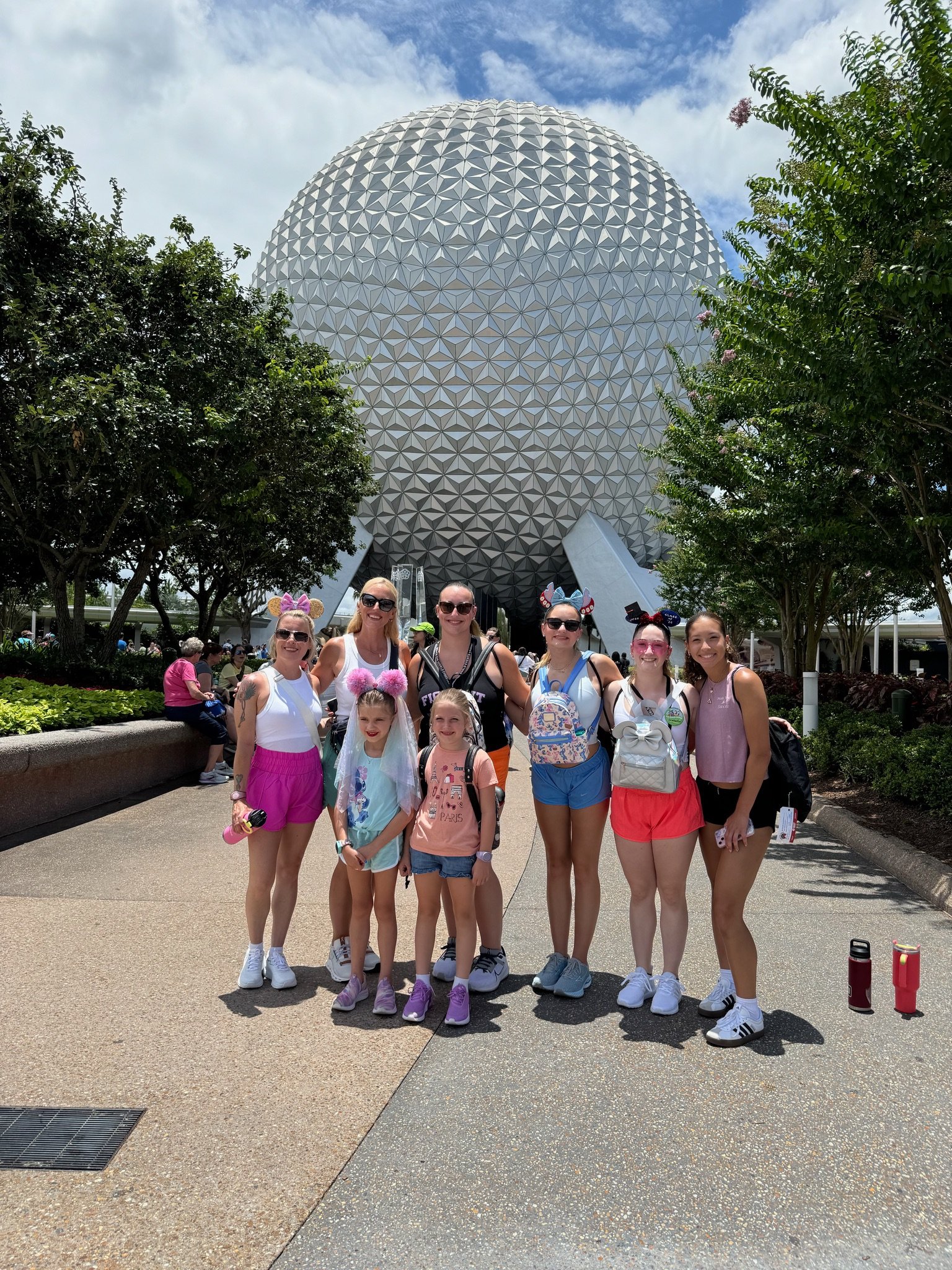 Group of nine people, including children, posing in front of the Epcot Spaceship Earth geodesic sphere at Disney World, Florida, on a sunny day with some trees and clouds in the background.