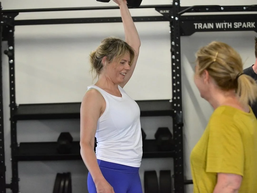 A woman in a white tank top and blue workout pants is lifting a weights above her head in a gym, smiling and interacting with another woman in a yellow shirt.