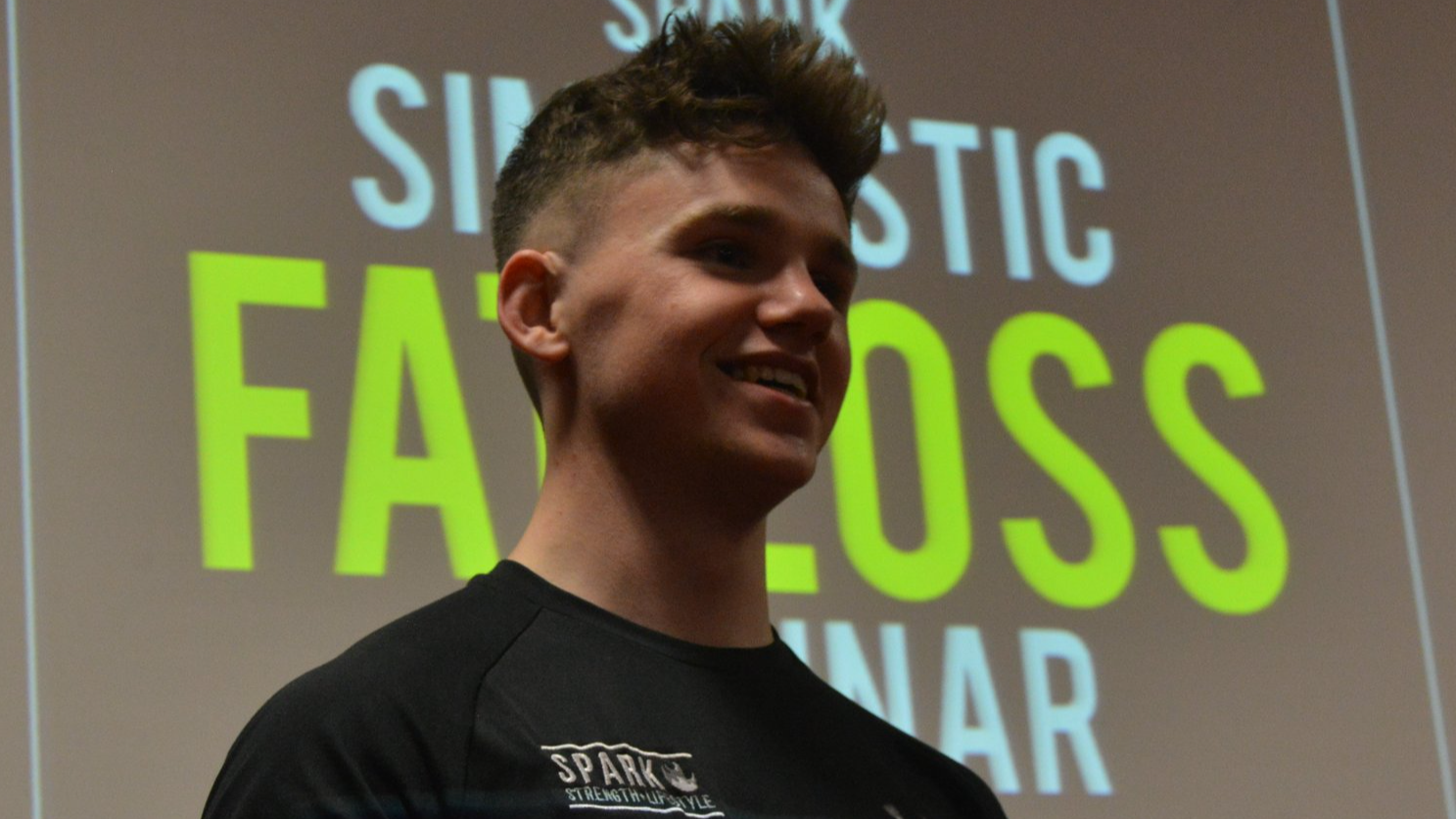 A young man with short, styled hair smiling in front of a projection with the words 'STATIC', 'FALLOS', and 'SEMINAR'.