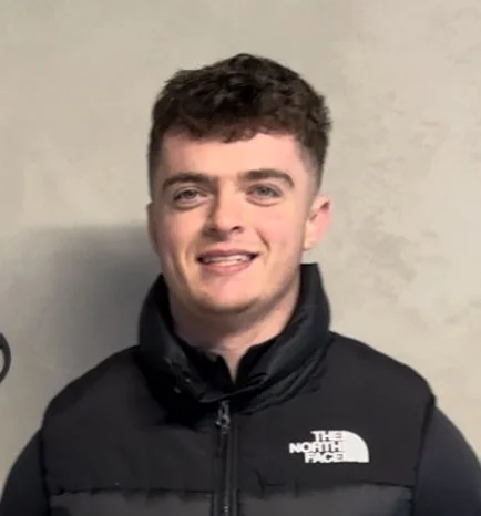 Young man with short curly brown hair smiling, wearing a black puffer vest with The North Face logo, standing against a plain beige wall.