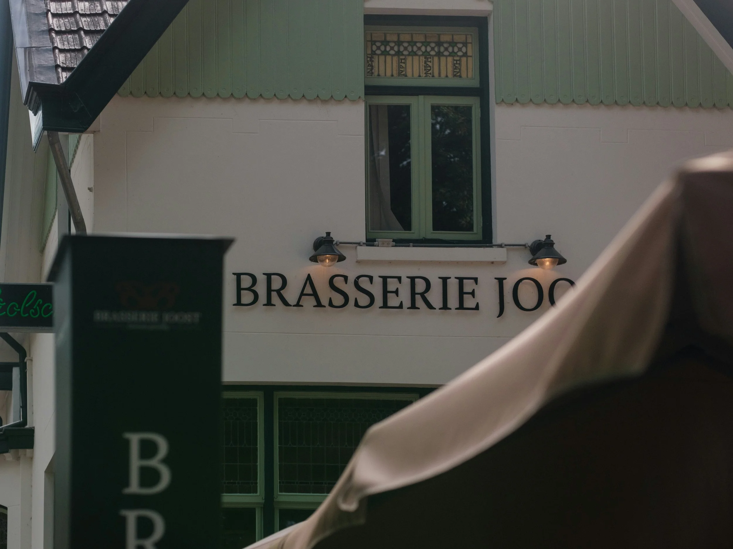 Extérieur d'une brasserie appelée 'Brasserie Jock' avec deux lampes au-dessus du nom, une fenêtre à l'étage et un parapluie beige en premier plan.