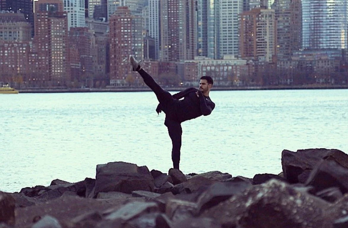 A man practicing a yoga pose on rocks near the water with a city skyline in the background.
