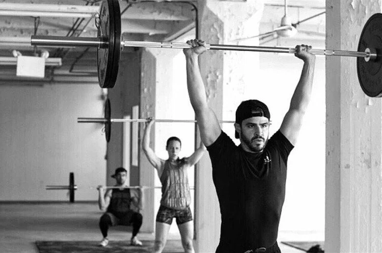 Three people lifting barbells in a gym, with the main person in the foreground and two others in the background.