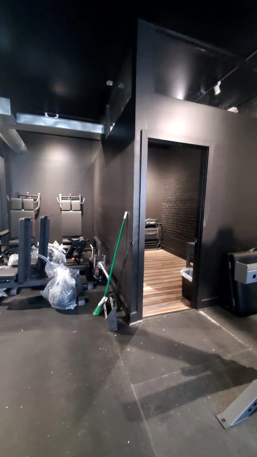 A room with exercise equipment, including two black workout machines and a chair, and cleaning supplies such as a broom, shovel, and plastic bag. There is a doorway to a small, dark room with stacked chairs and a trash bin nearby.