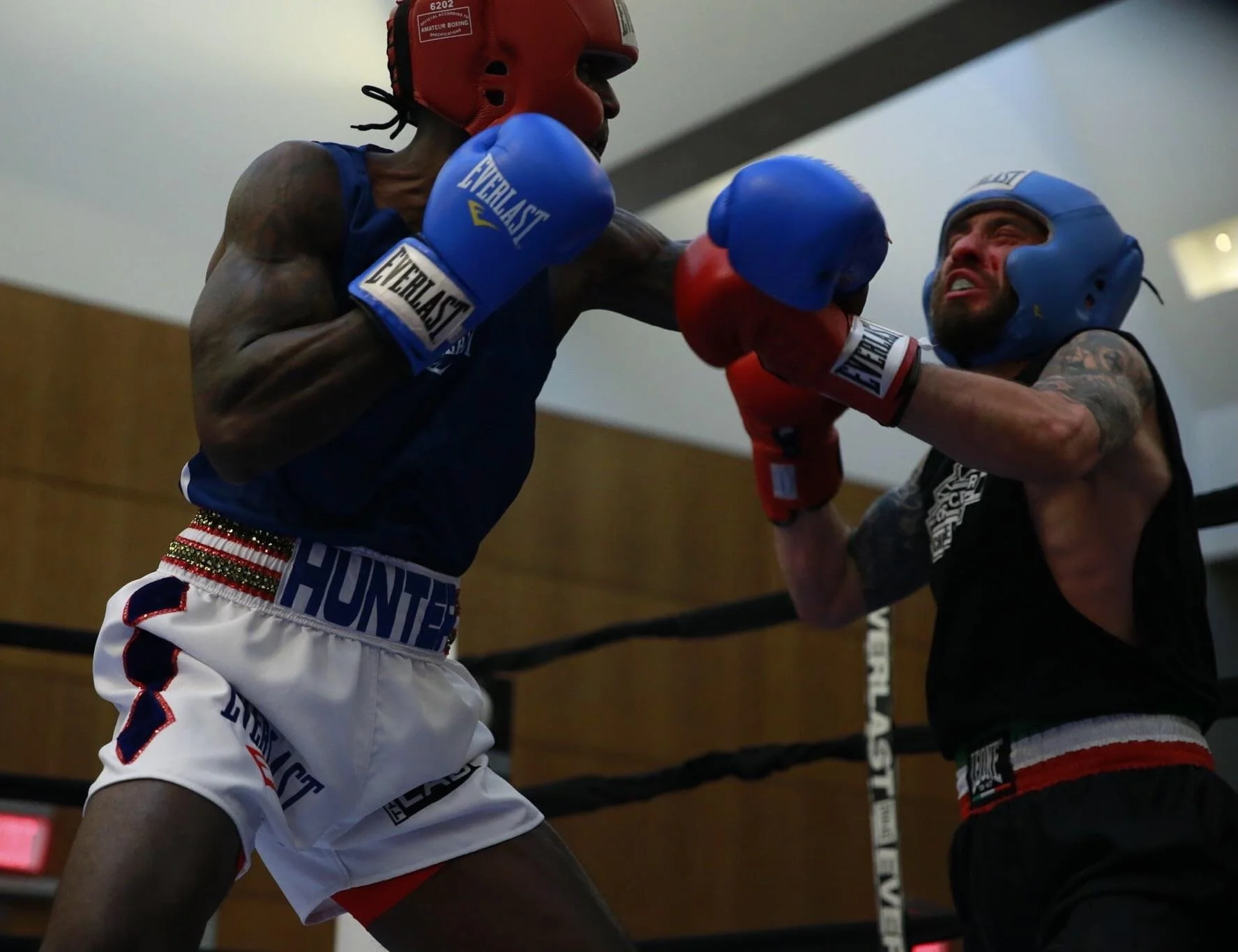 Two male boxers sparring in a boxing ring, one wearing red sparring gloves and red headgear, the other wearing blue gloves and blue headgear, with the boxer in red throwing a punch.