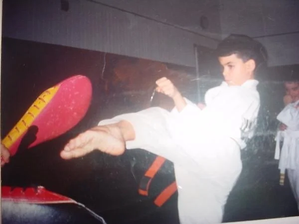 A young boy in a white martial arts uniform kicking a red padded target held by someone else, indoor setting, another person in martial arts uniform in the background.