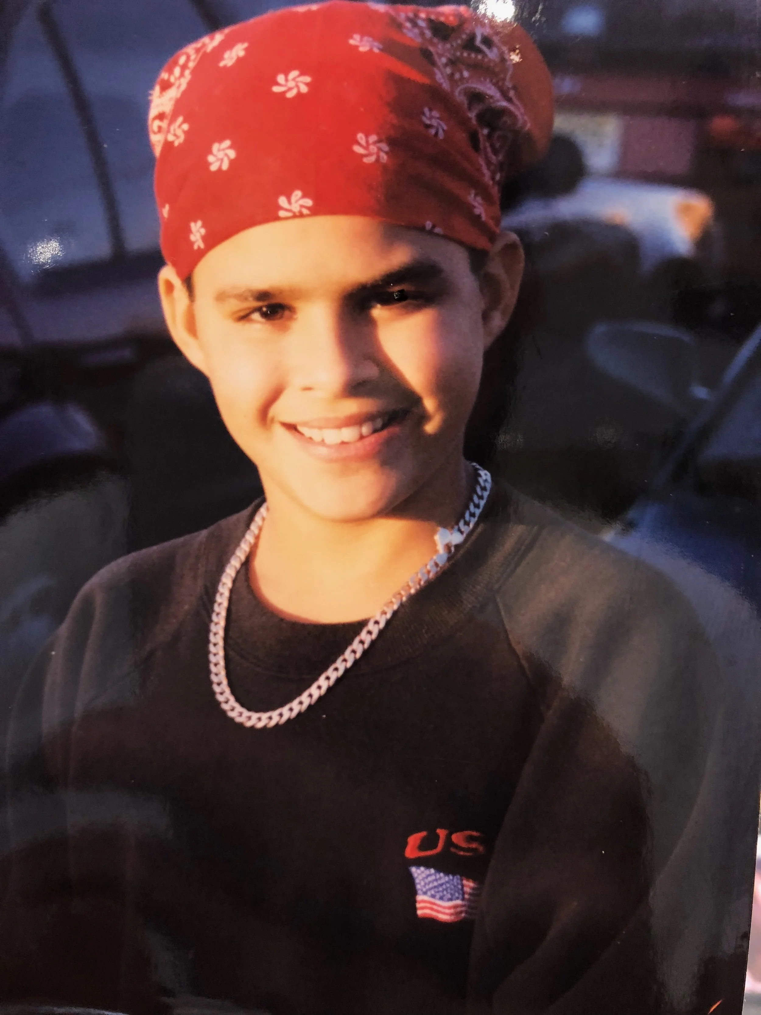A young man smiling, wearing a red bandana with white flower patterns, a black sweatshirt with a US flag patch, and a thick chain necklace, sitting in an indoor setting.