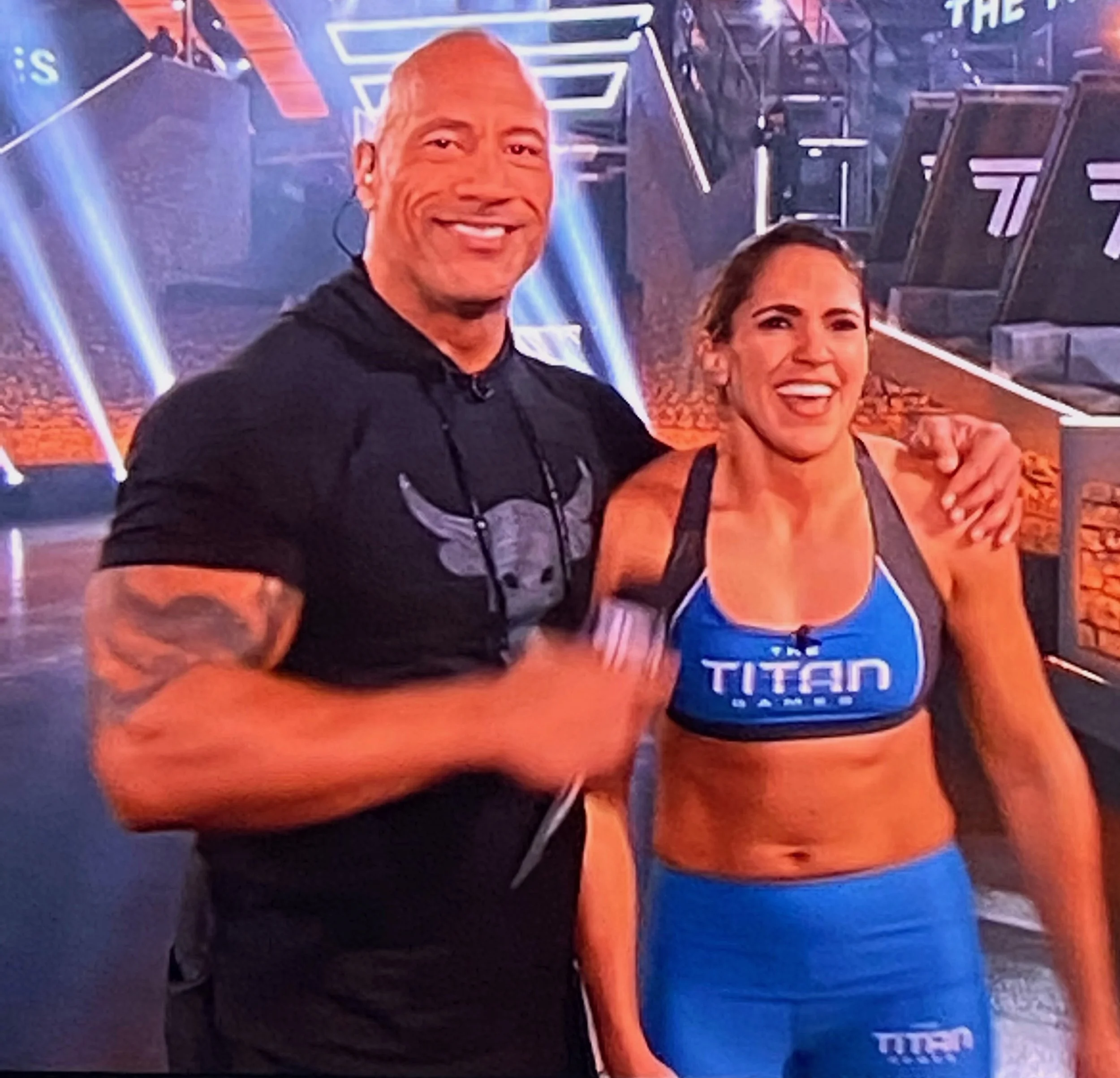 A man and a woman smiling and posing together in a gym or fitness studio. The man is wearing a black T-shirt with a bull logo and has tattoos on his arm. The woman is wearing a blue sports bra and matching blue shorts with the words "TITAN" on them. The background features gym equipment and colorful lights.