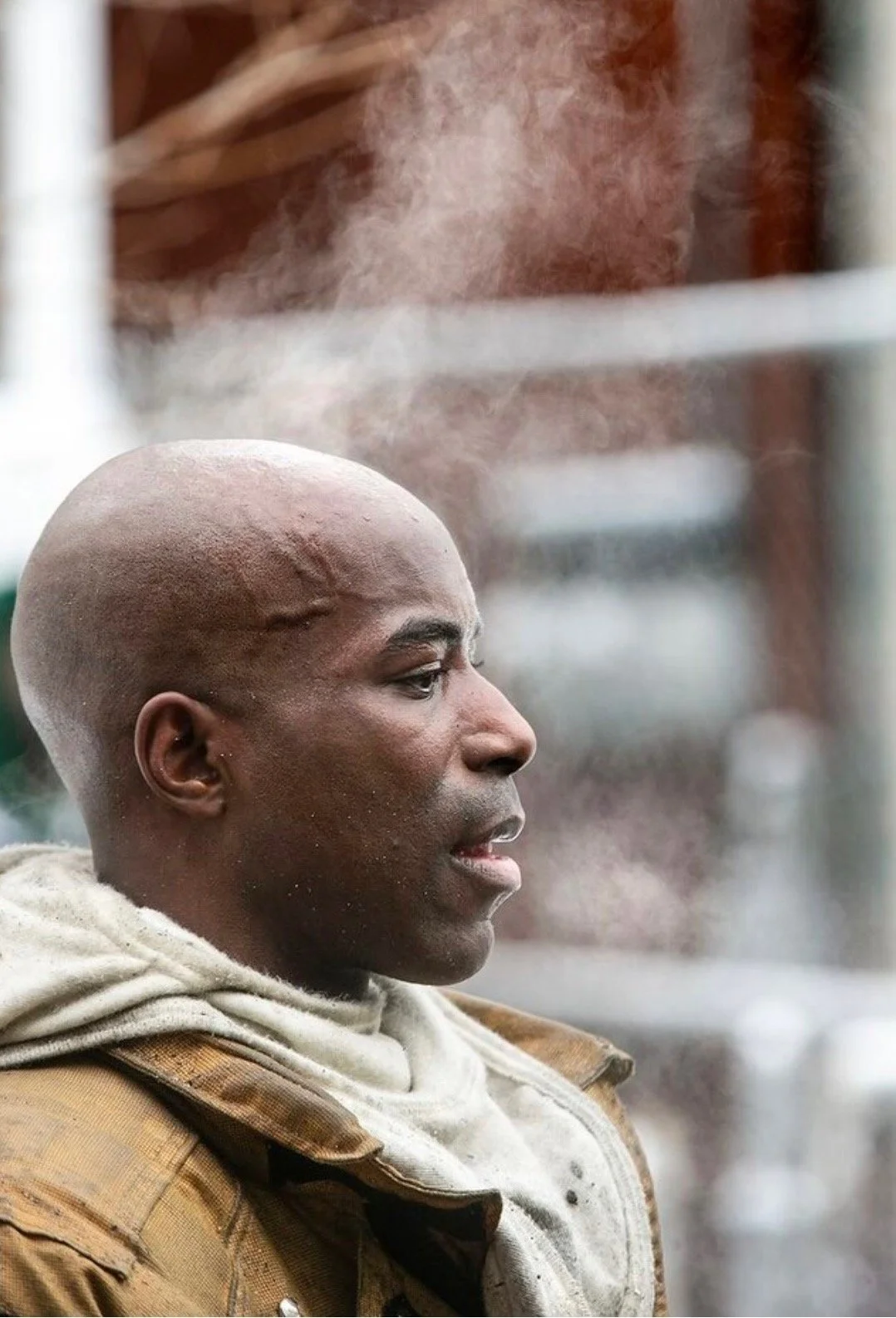 A man with a shaved head and wearing layered clothing stands in cold weather, with visible breath vapor in the air.