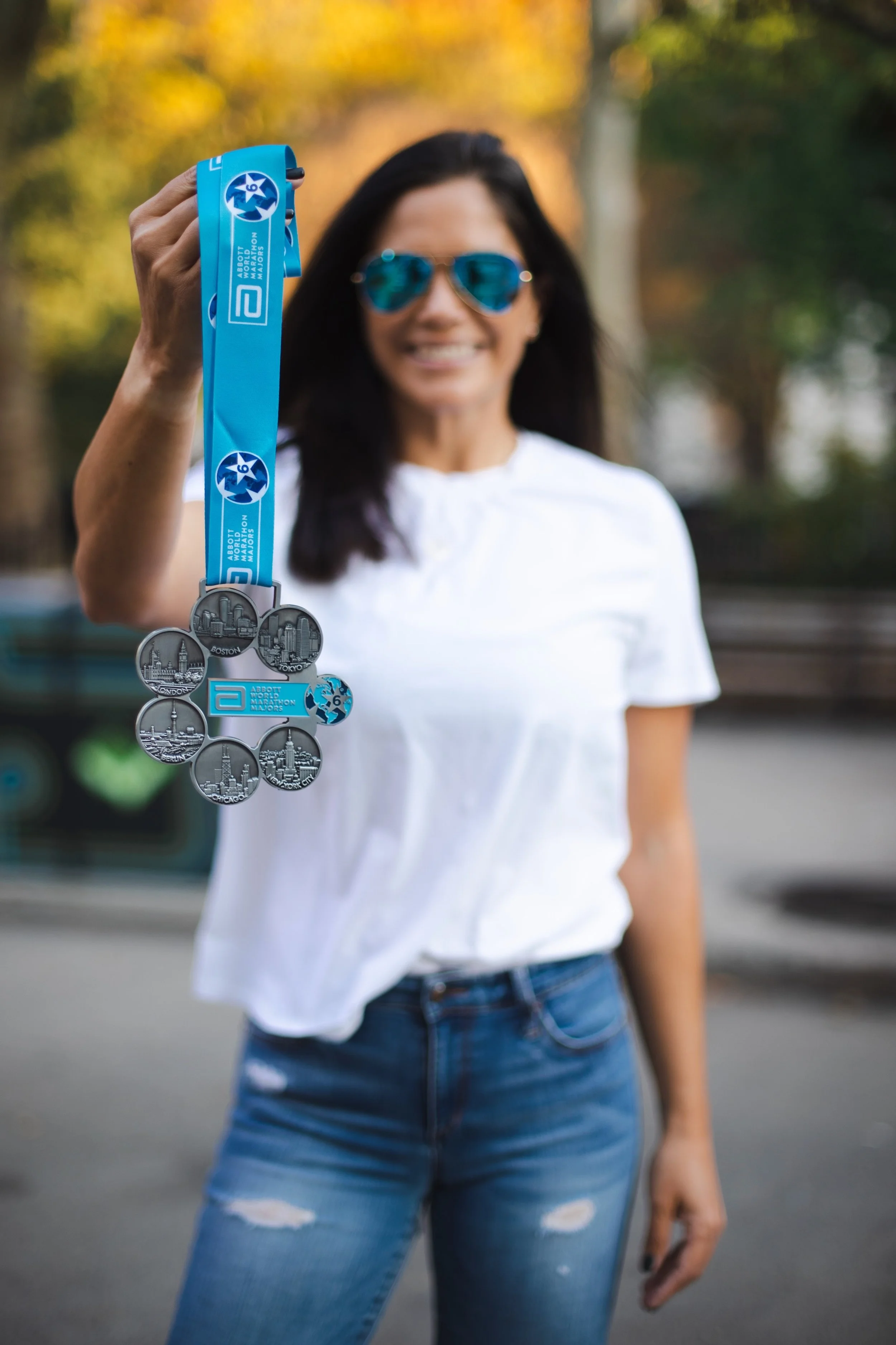A woman in a white shirt and blue jeans holding up a marathon medal with a blue ribbon, outside with trees in the background.