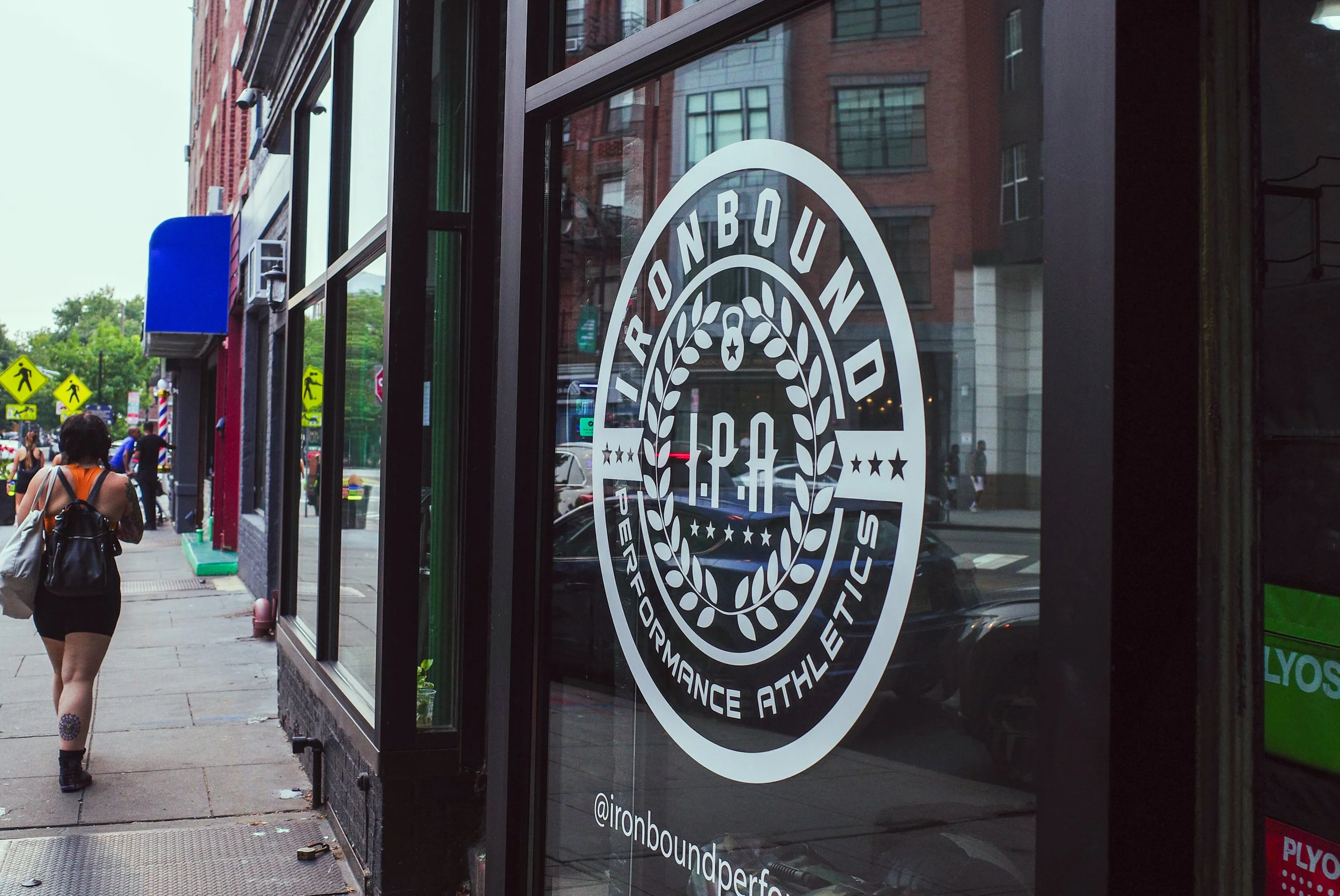Storefront window with Ironbound Performance Athletics logo, a black and white circular emblem with the words 'Ironbound Performance Athletics' and a wreath design, on a city sidewalk with pedestrians and buildings in the background.