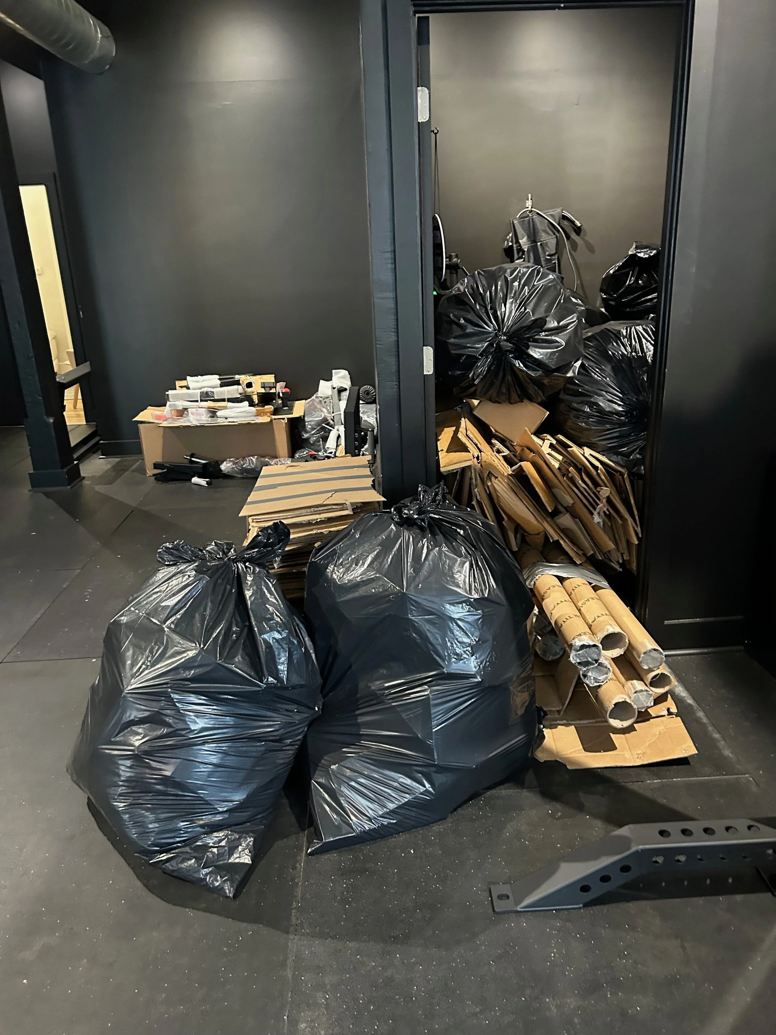 Black trash bags, cardboard, and paper tubes stacked near a black wall in an indoor space.