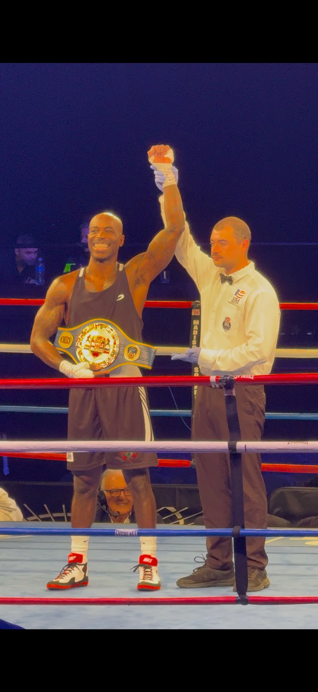 A victorious boxer in a boxing ring, wearing a championship belt, raising his right fist in celebration, with a referee holding his hand. The boxer is smiling, dressed in athletic gear, with a background of spectators and a dark environment.