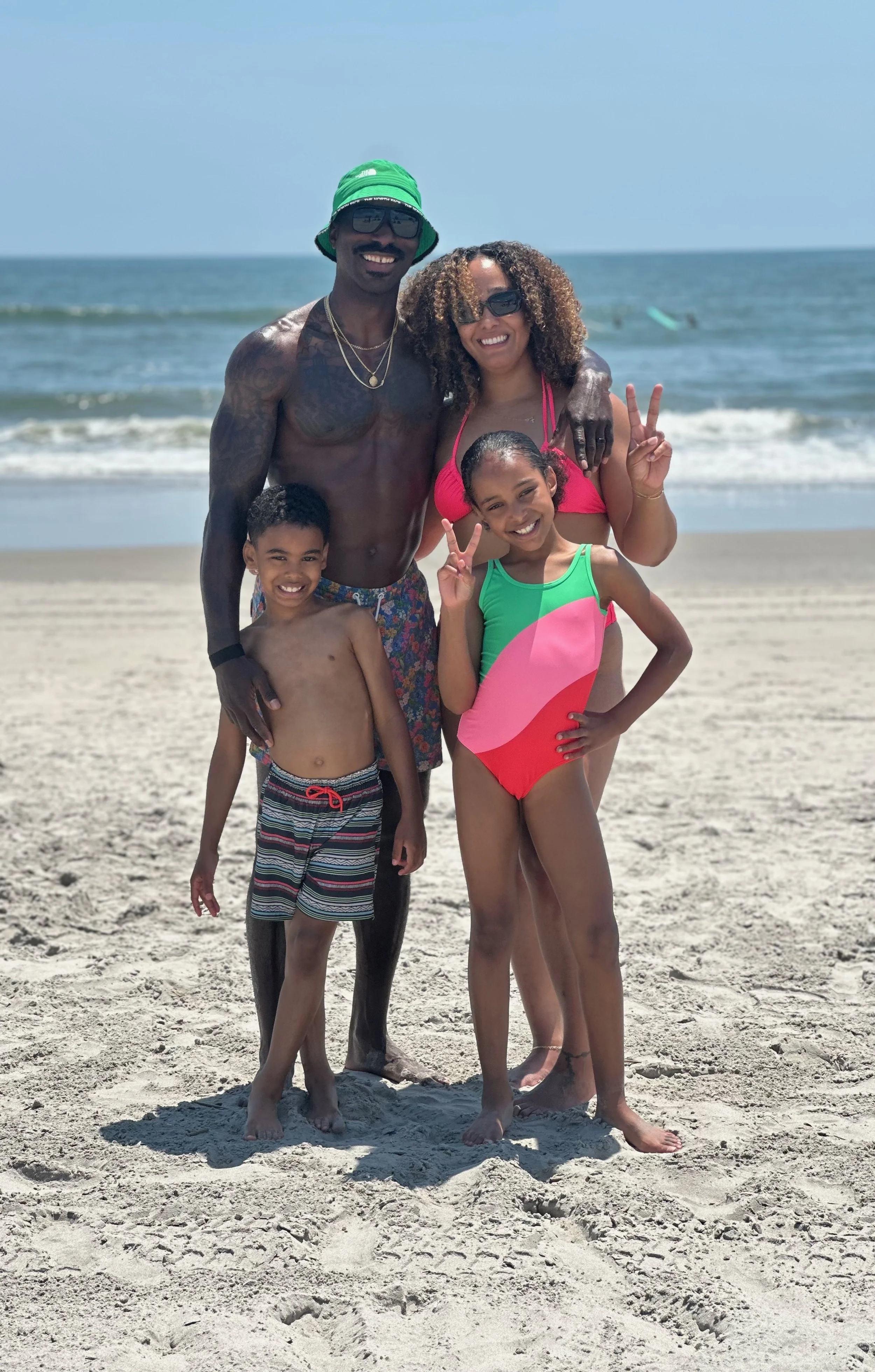 A family of four smiling on the beach with the ocean in the background. The father and mother are standing behind their young son and daughter, all in swimwear and sunglasses, making peace signs.