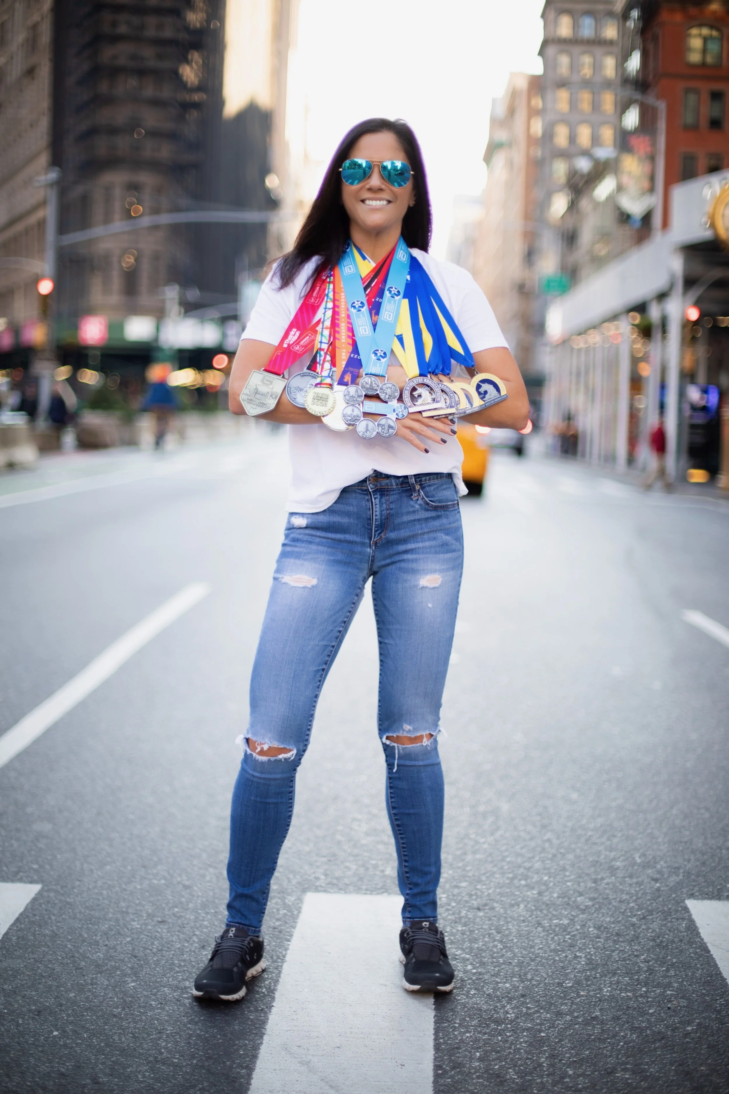 A woman standing in the middle of a city street, wearing sunglasses and a white T-shirt, displaying an array of medals around her neck, with some medals hanging from her hands.