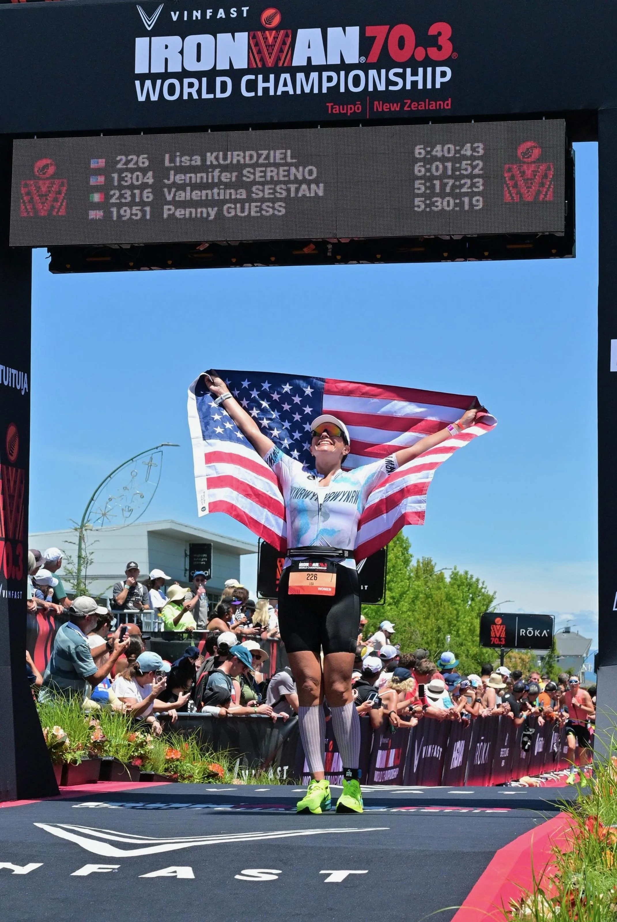 Female marathon runner at the finish line of the Ironman 70.3 World Championship in Taupō, New Zealand, holding an American flag and smiling. The crowd cheers behind her and the race results display above.