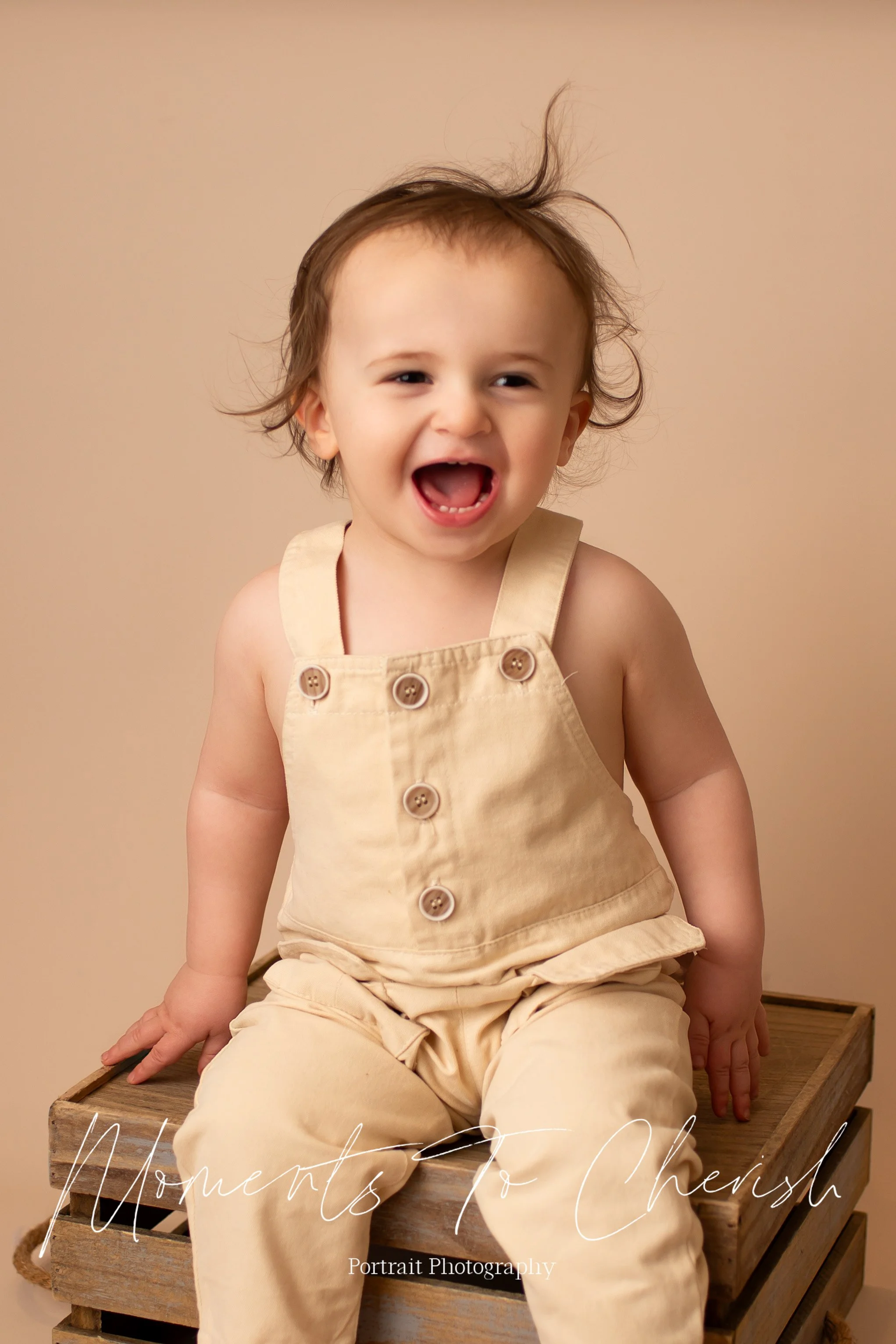 Toddler boy sitting on a wooden crate on a beige backdrop. He is wearing cream overalls and has an expression of excitement