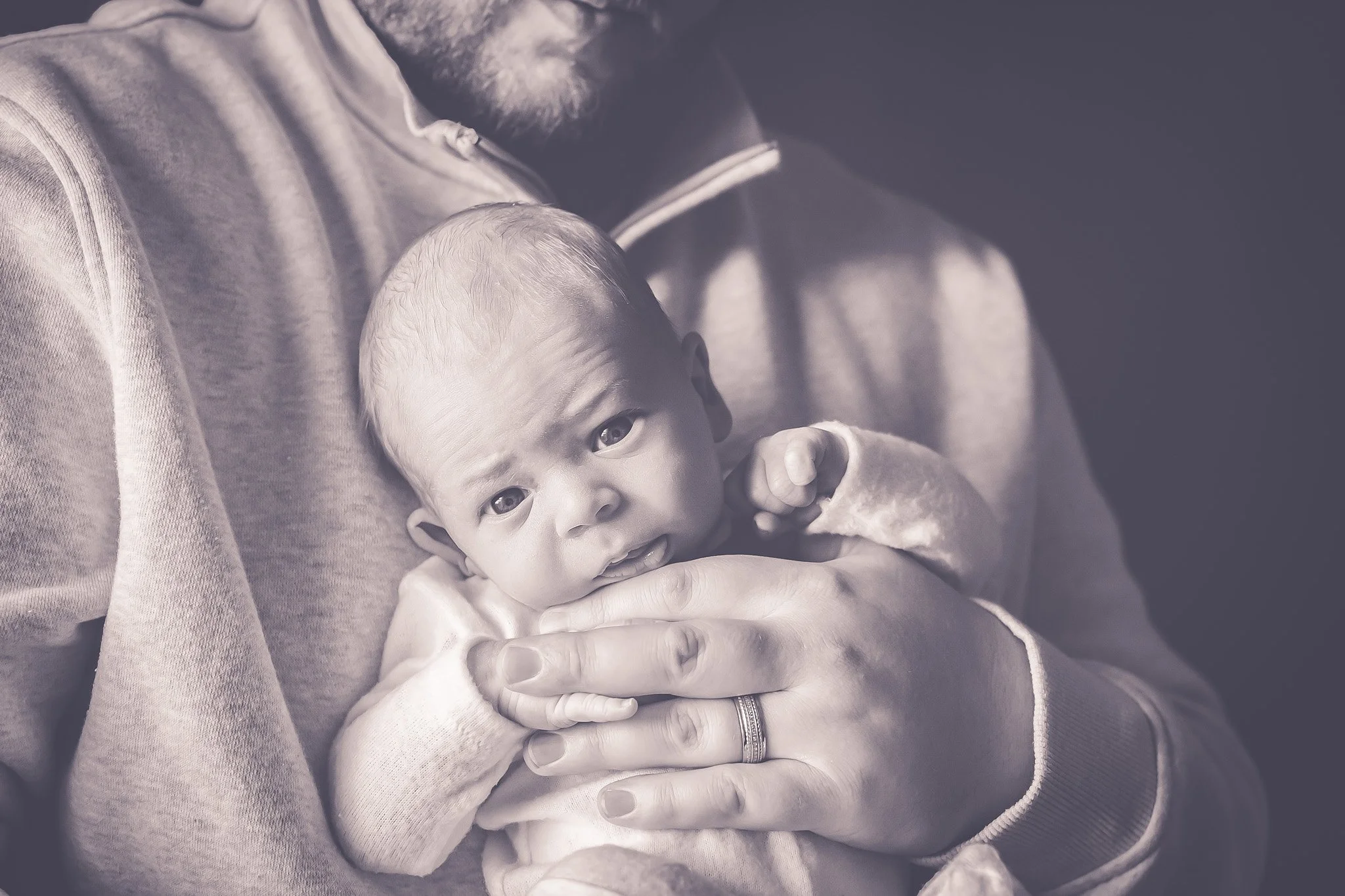 Black and white image of a close up newborn being held by dad. Only dad's hand and chin are visible. Baby is looking right into the camera.