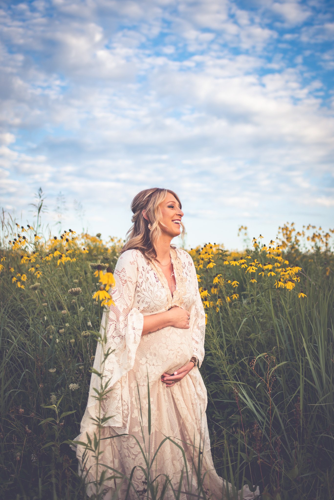 A pregnant woman in a white lace dress standing in a field of yellow flowers, smiling and holding her baby bump, with a blue sky and some clouds above.