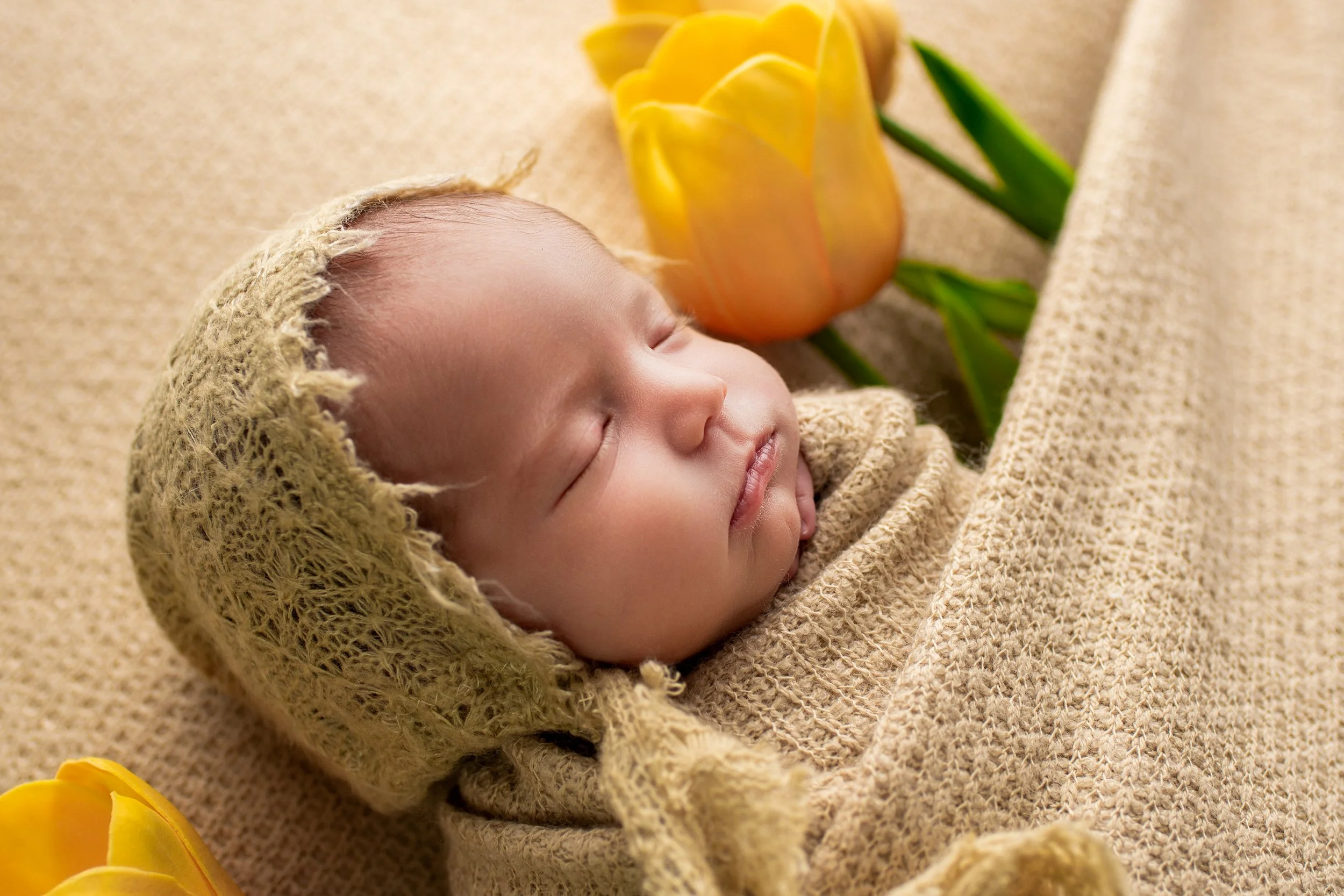 Close up of newborn baby girl wearing a bonnet and tucked in with yellow tulips