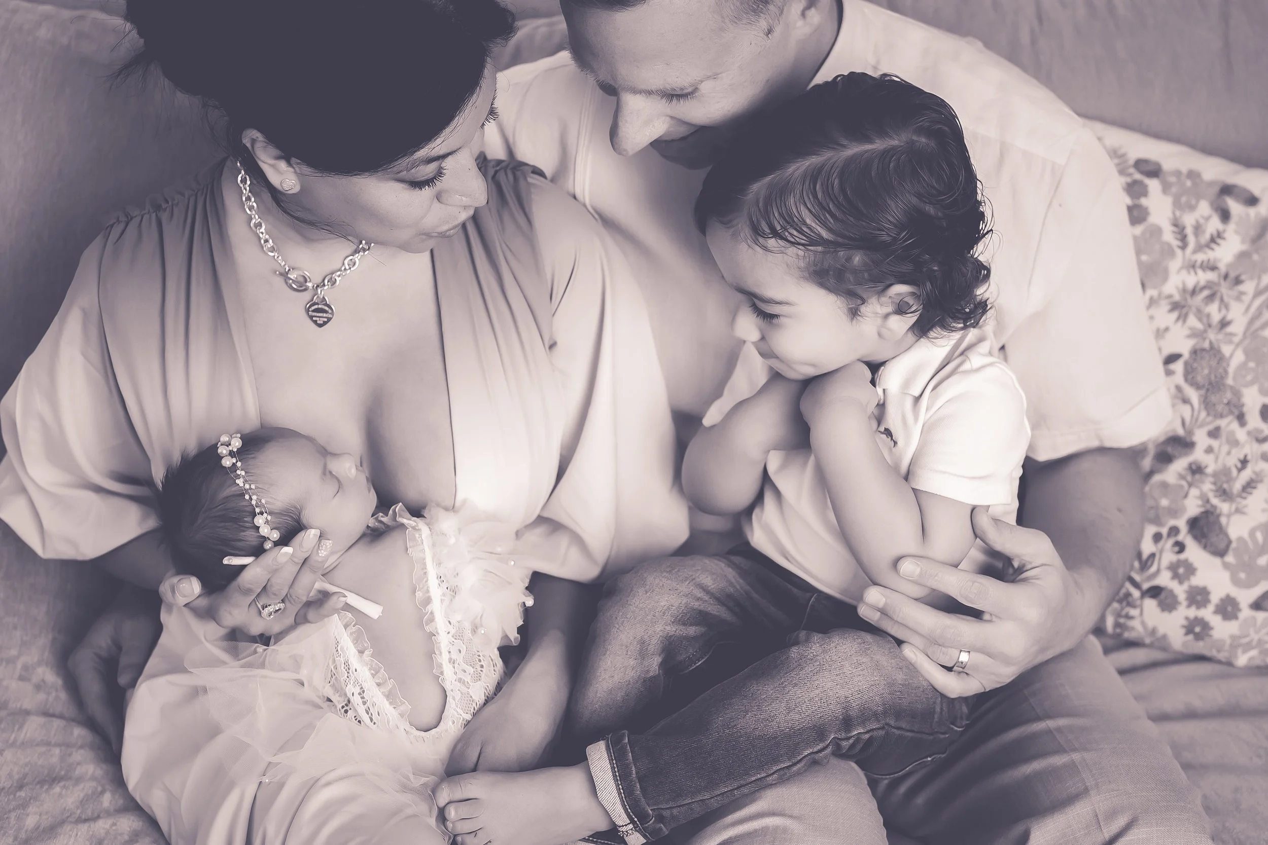 Black and white image of a family of four snuggling on the couch. Mom is holding a newborn baby girl that is wearing a lacey outfit. Dad is holding a toddler boy that is wearing a simple light polo shirt and jeans. Everyone is looking down at baby