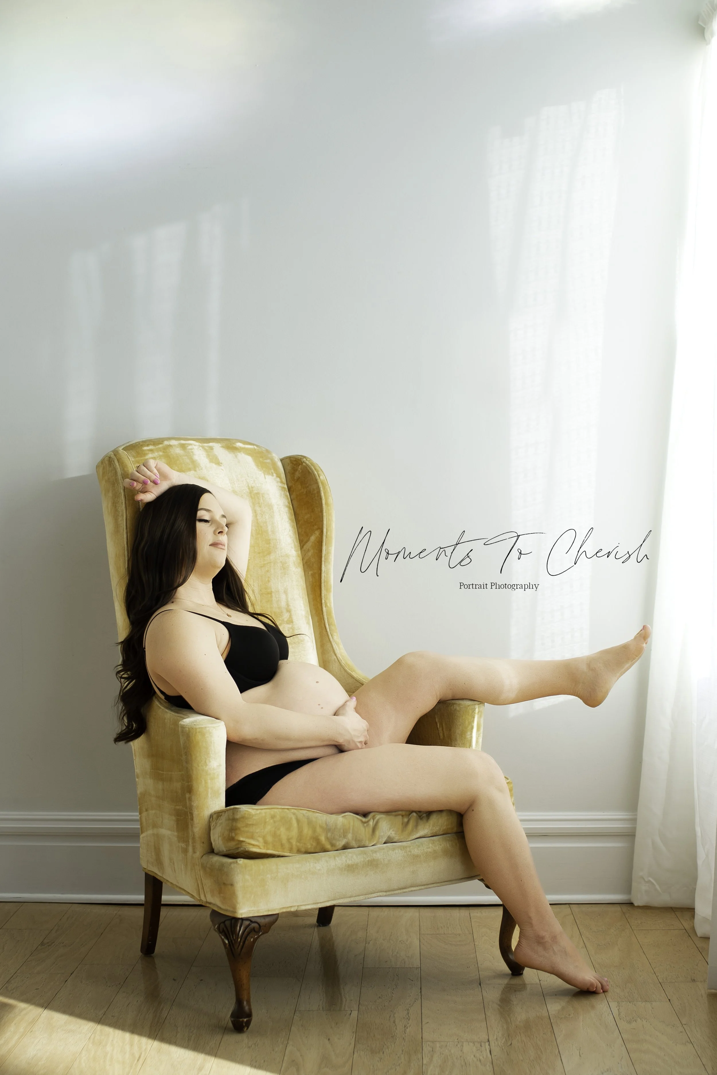 A pregnant woman with long dark hair reclining in a yellow velvet armchair, wearing black lingerie, in a bright room with hardwood flooring and white curtains. She has her eyes closed and one hand resting on her forehead, with one leg extended and re