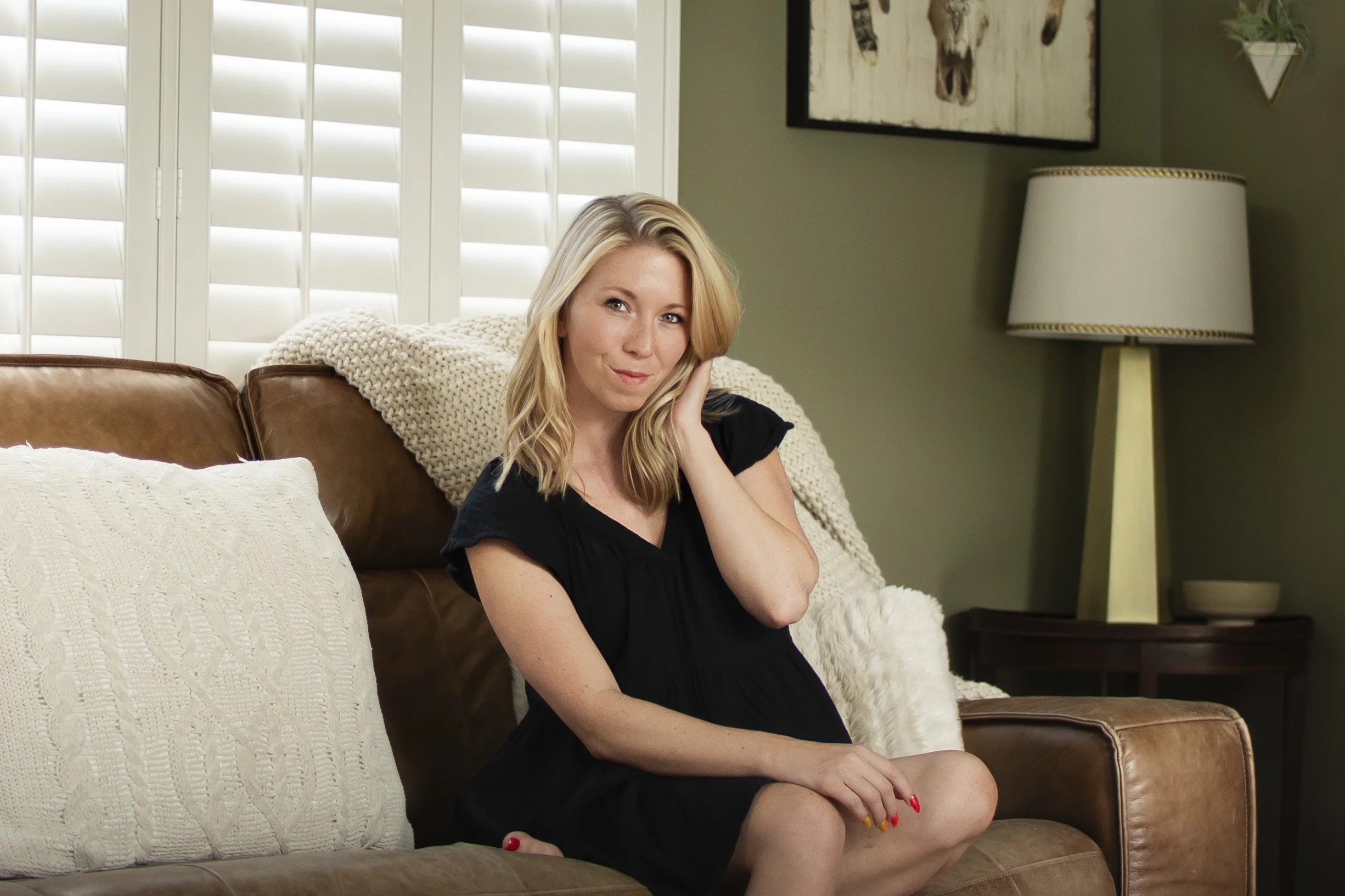 A woman with blonde hair wearing a black dress sitting on a brown leather couch in a living room, with one hand resting on her cheek and a subtle smile.