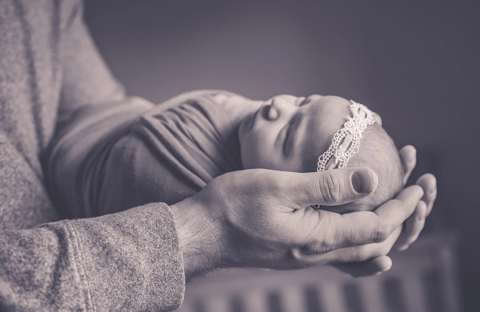 Black and white image of a newborn baby girl being held in her dad's hands. She has a lacey headband on and is swaddled.