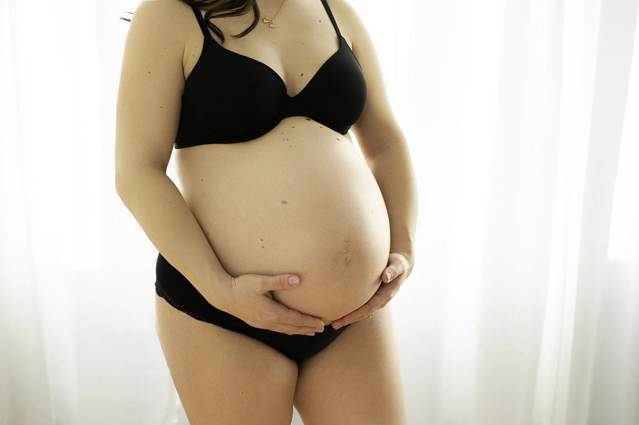 A pregnant woman is standing with her hands gently placed on her belly, wearing black lingerie, in a bright room with white curtains.