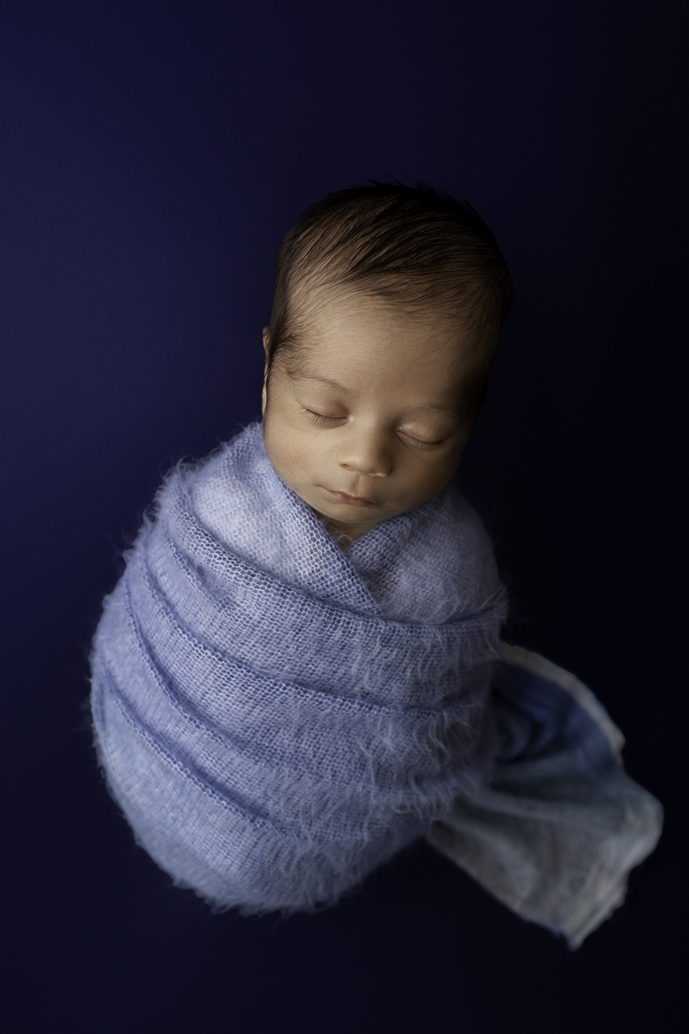 Newborn baby boy wrapped in a light blue swaddle on a dark blue backdrop 