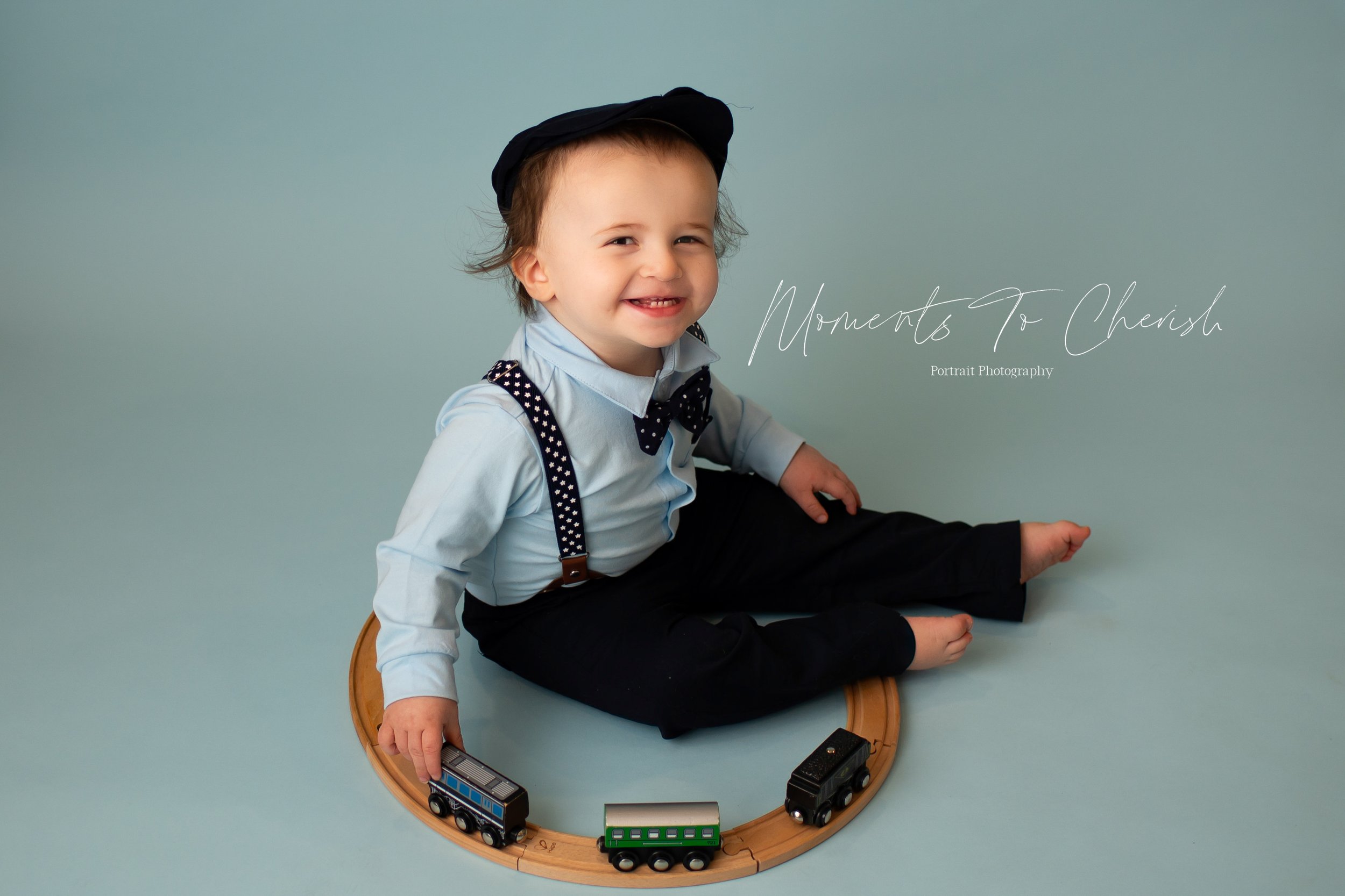 Toddler boy with a newsboy style hat on. He is sitting on the floor and looking up towards the camera. He has a bowtie and suspenders on along with dress pants and a dress shirt. He is on a blue background and playing with a wooden train set
