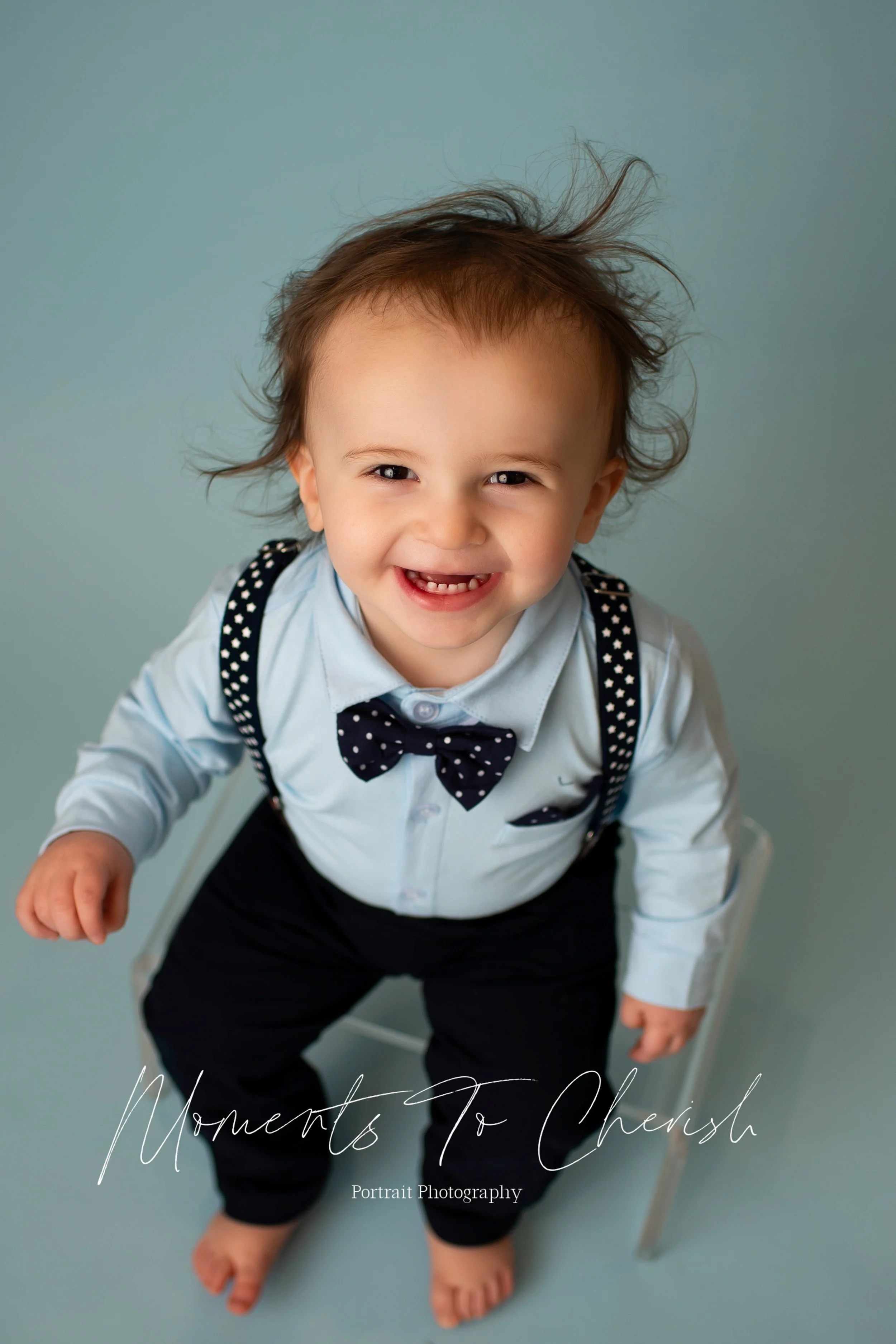 Toddler boy wearing suspenders and a bow tie. He is looking up and smiling. He is on a blue backdrop.