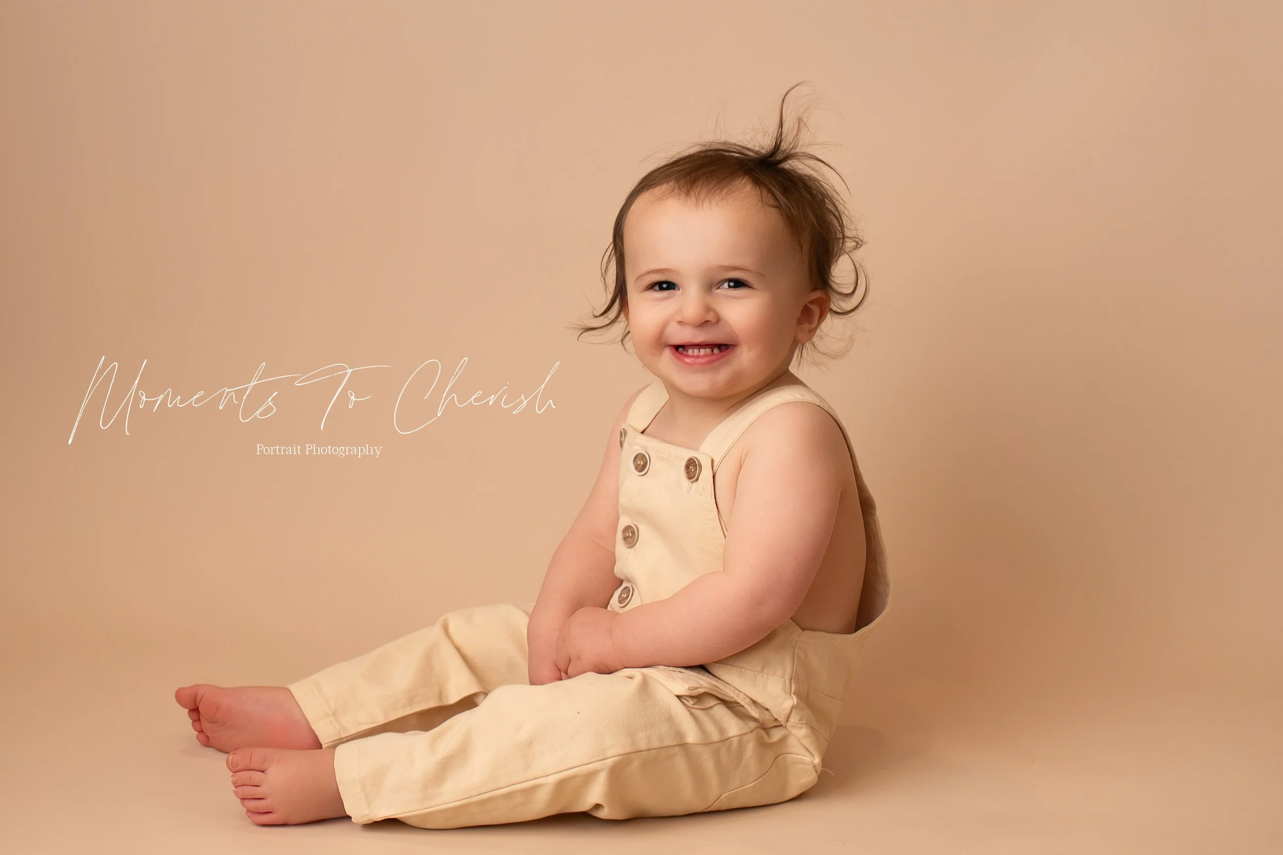 Toddler boy sitting on the floor on a beige background. He is wearing cream colored overalls and has a big smile on his face