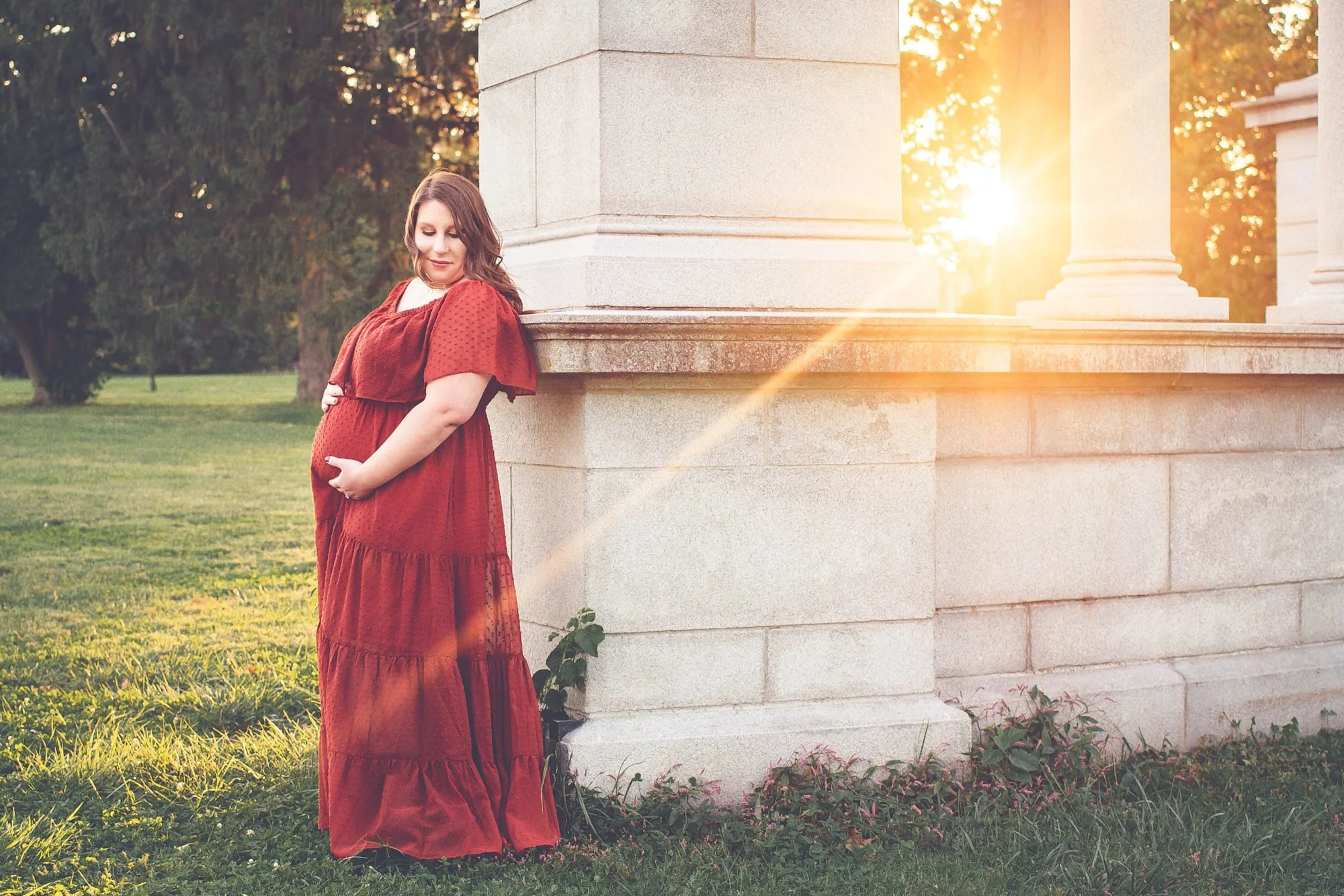A pregnant woman in a long red dress leaning against a stone structure at sunset in a park.