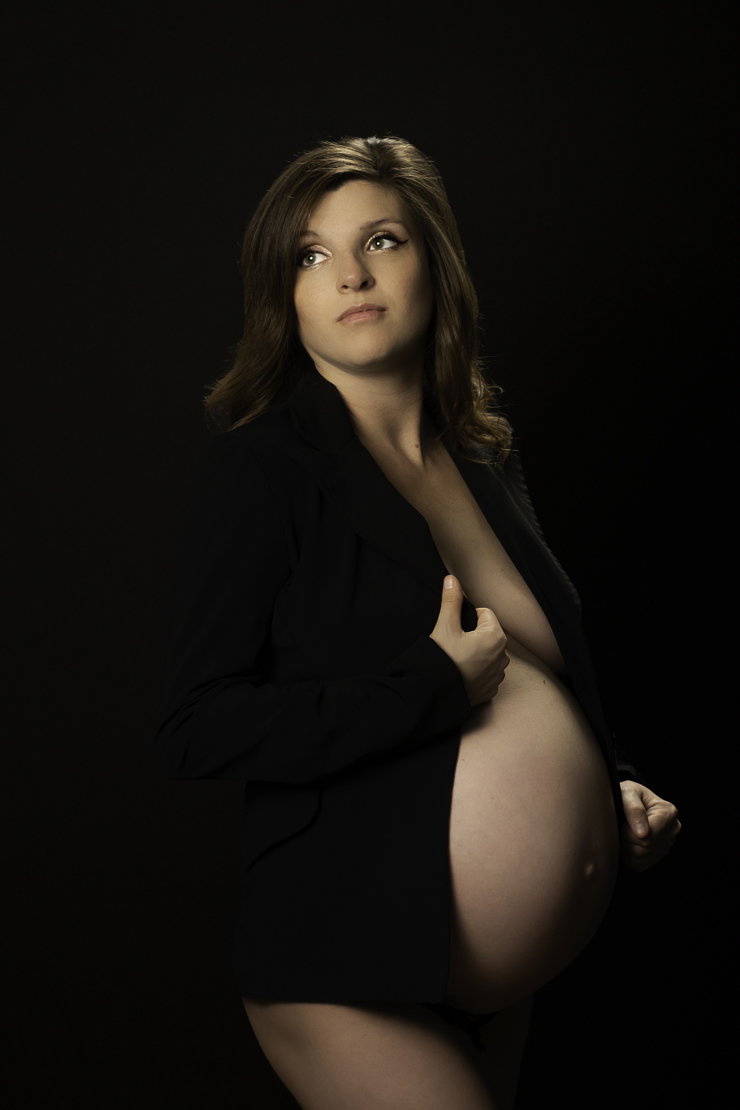 A pregnant woman in a black blazer holding her belly against a dark background.