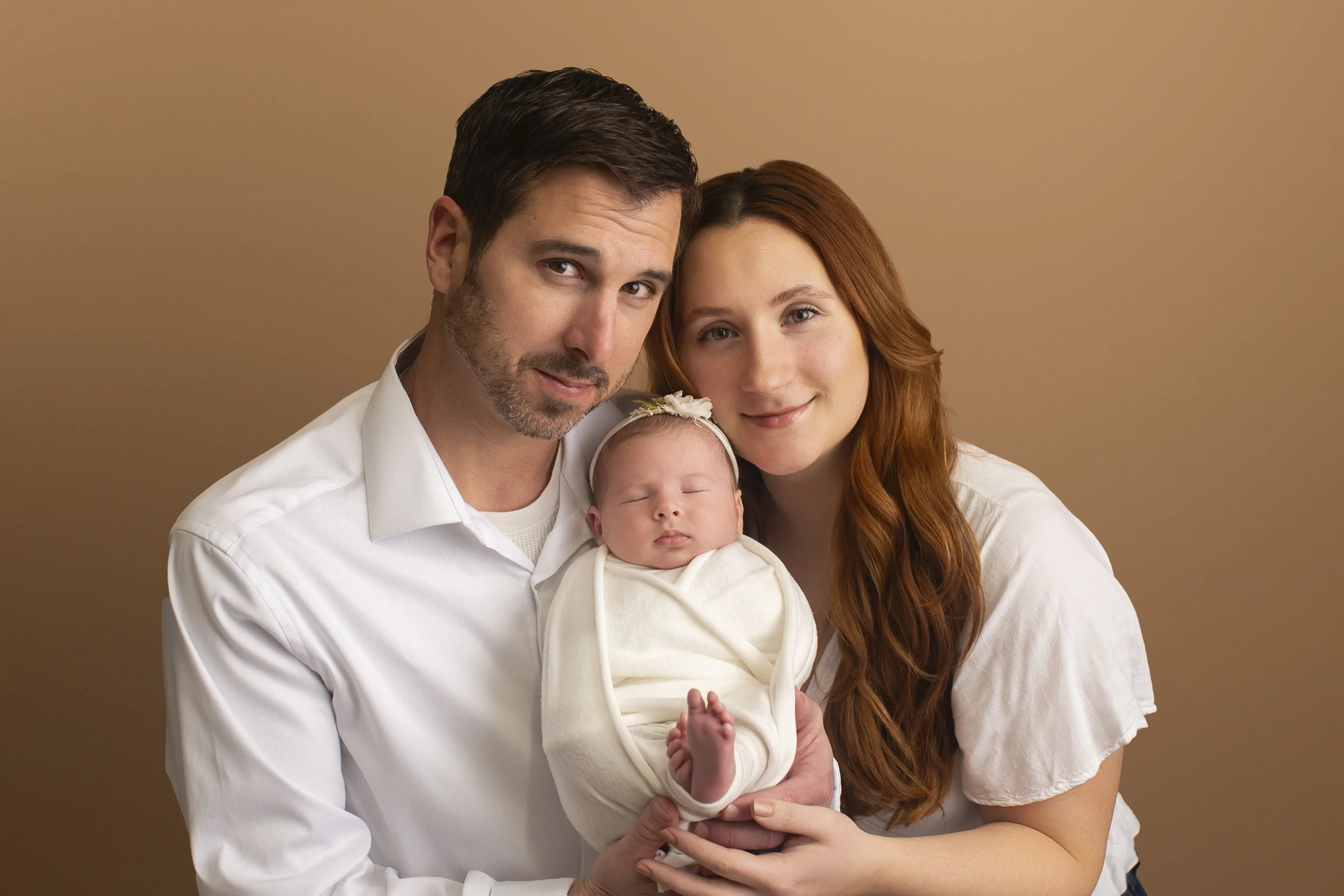 Close up family photo with mom and dad wearing white shirts holding a newborn baby girl that is in a white swaddle. They are on a mocha colored backdrop