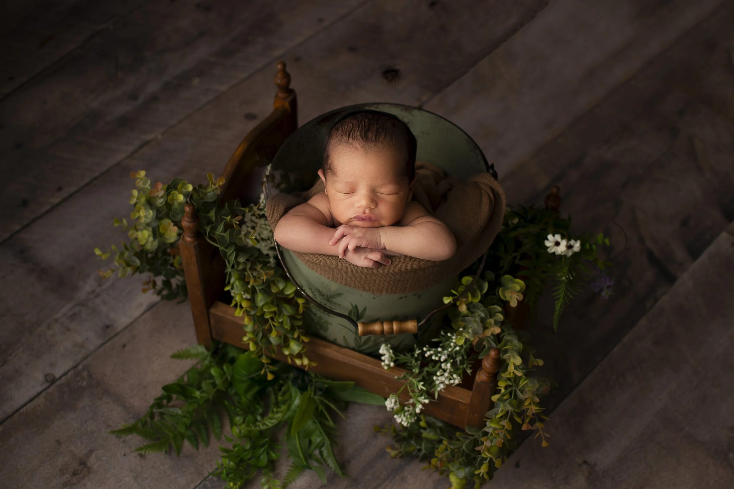 Newborn baby boy laying on his hands in a bucket surrounded by greenery