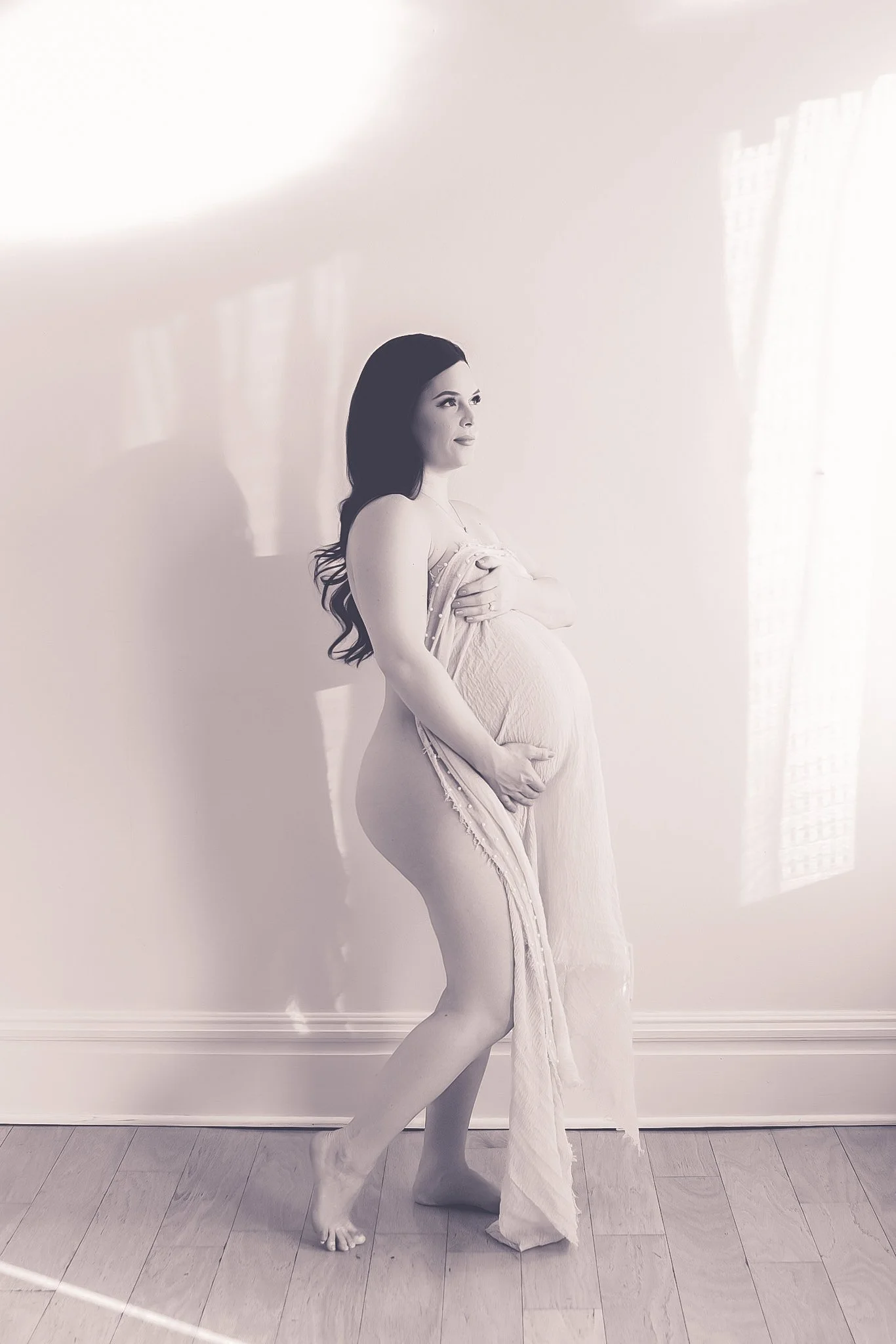 A pregnant woman with long dark hair standing barefoot on a wooden floor, holding a sheer fabric in front of her chest, gazing forward, with sunlight streaming through a window in the background.