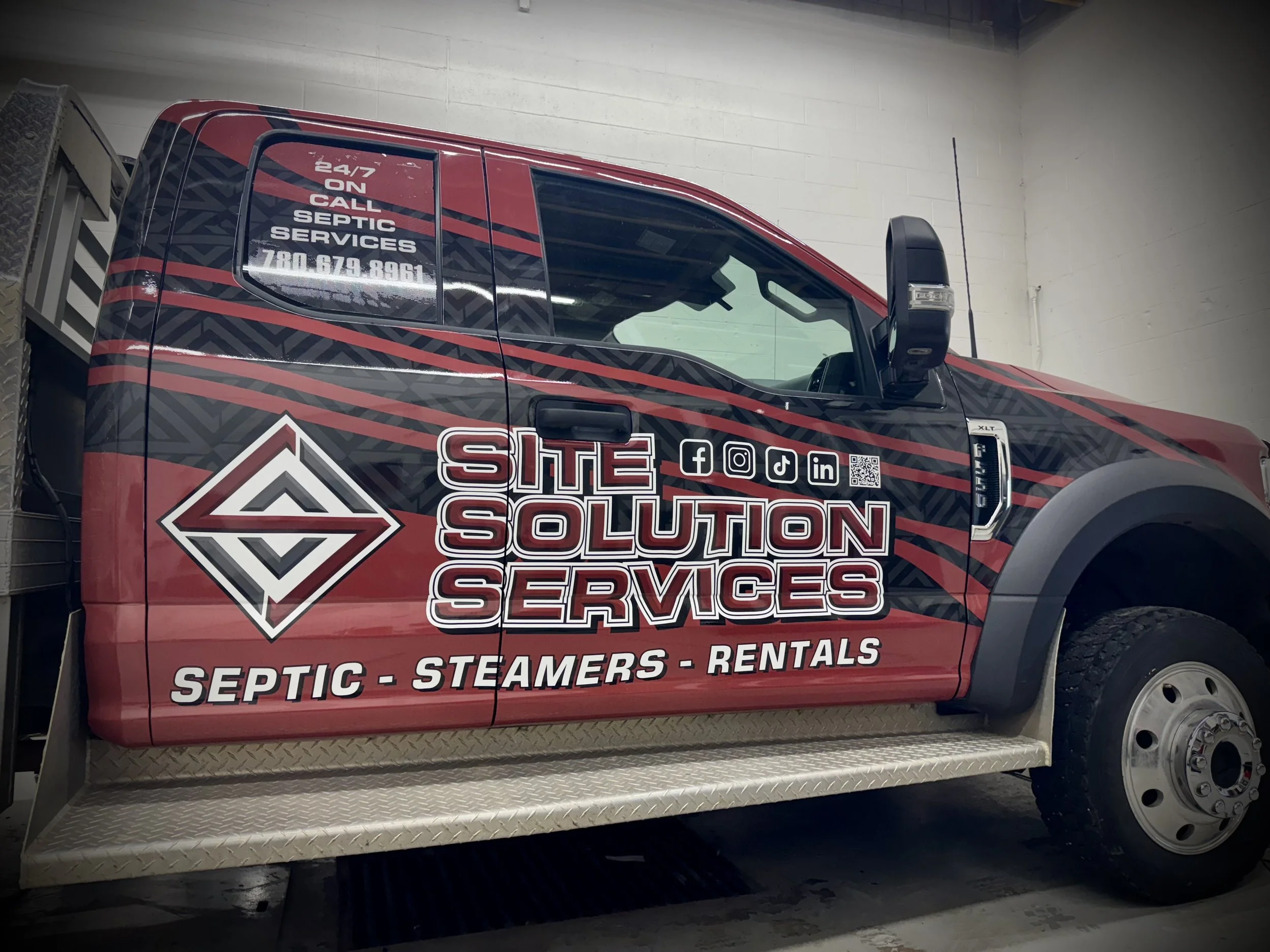 A fleet of four construction and service vehicles with matching company logos and branding traveling along a rural road at sunset, including a white pickup truck, a white semi-truck, a red dump truck, and a tracked excavator.