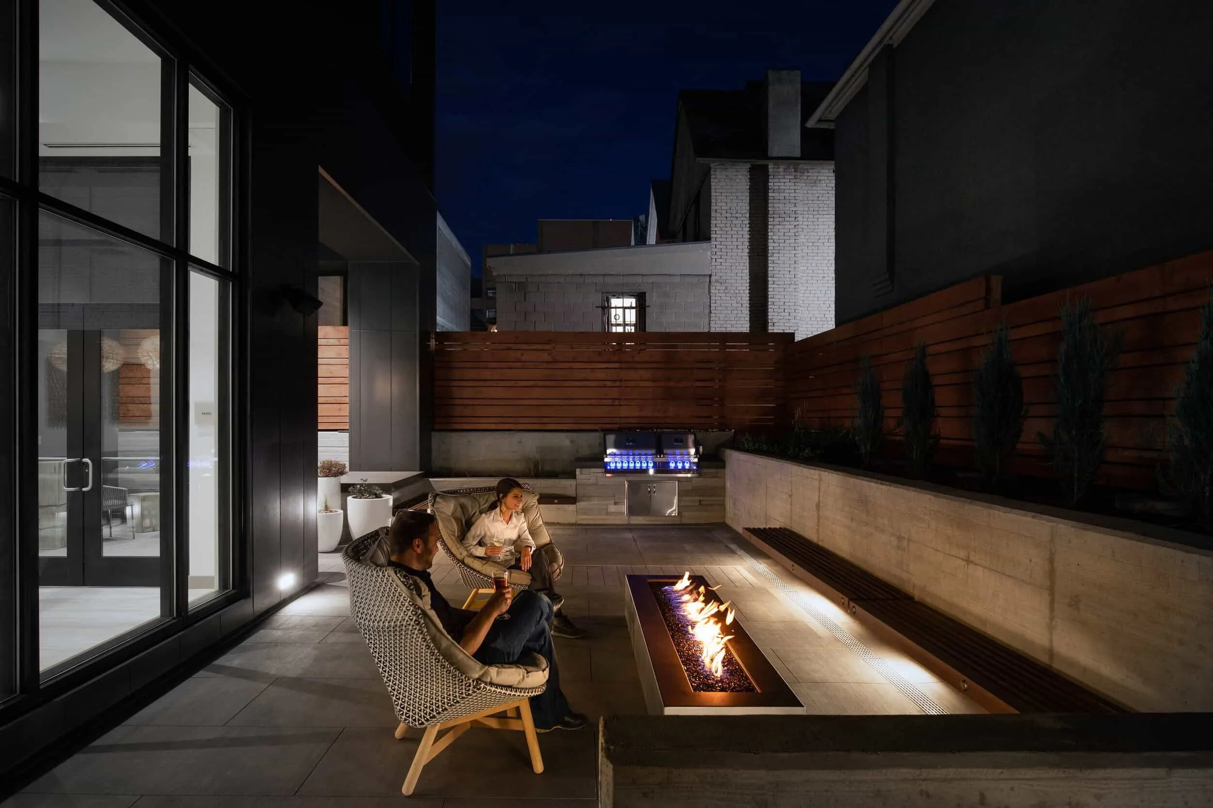 Two people are sitting on outdoor patio chairs around a fire pit at night. There is a grill in the background and wooden fencing enclosing the space. The area is decorated with plants and modern architecture.