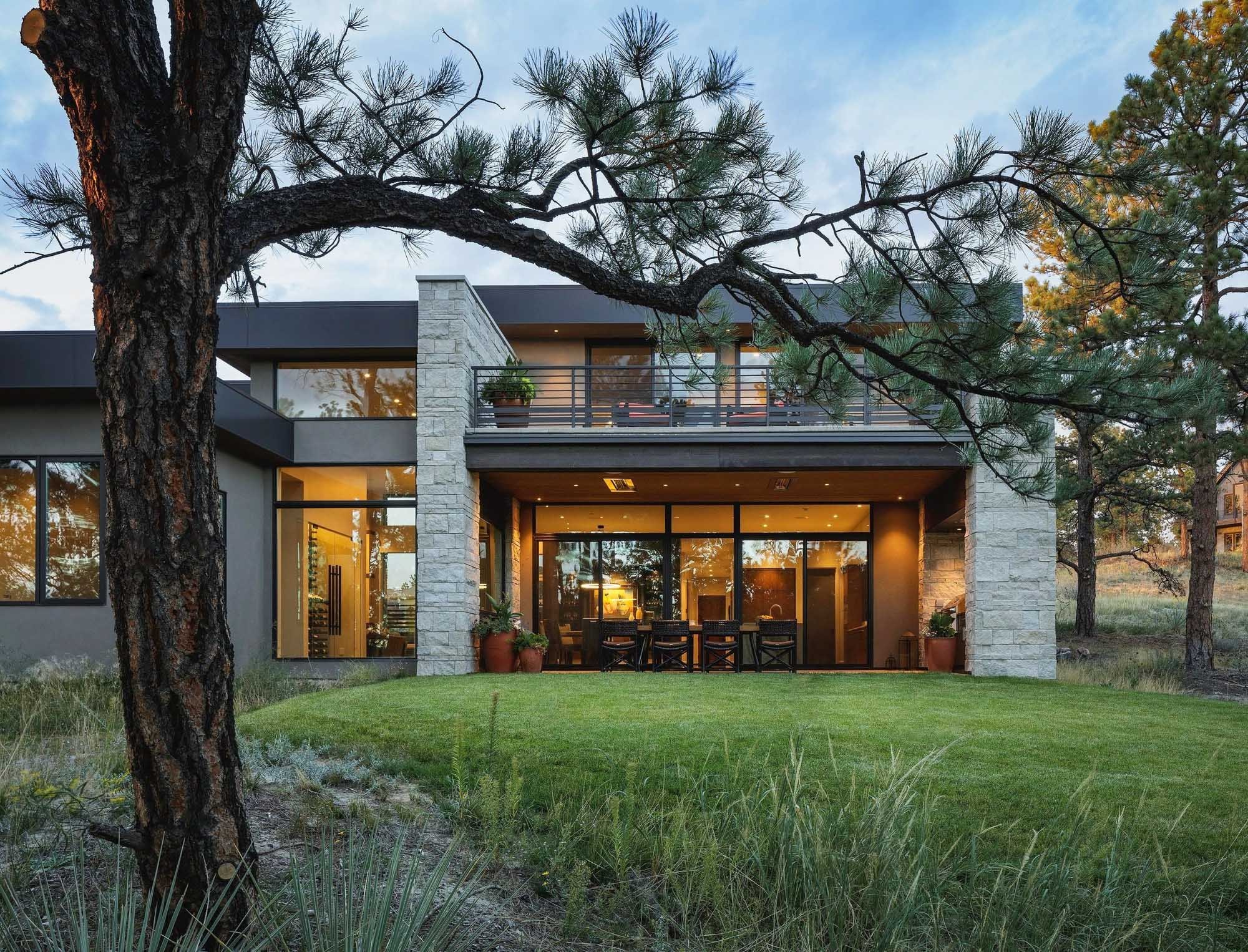 A modern house with large glass windows and stone accents, surrounded by trees and a well-manicured lawn, during sunset.