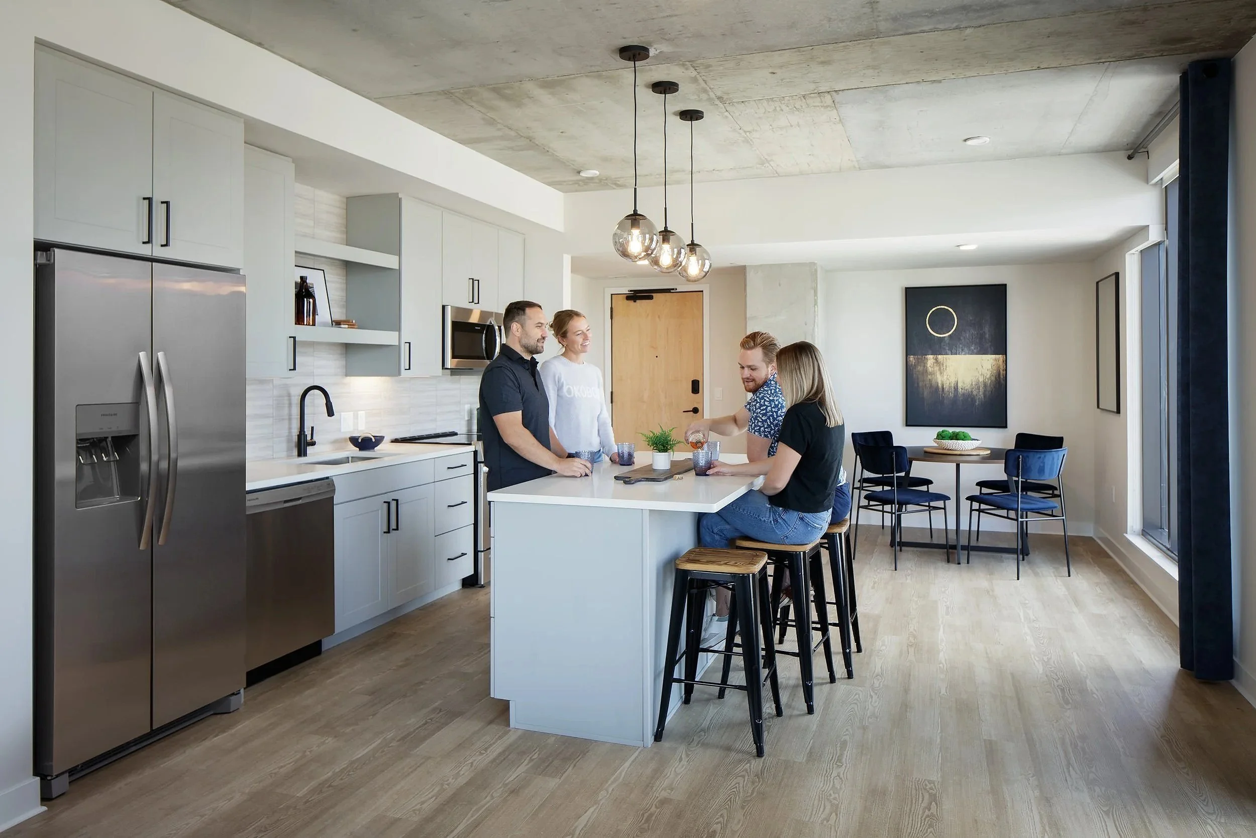 Four people gathering in a modern kitchen with white cabinets, a stainless steel refrigerator, and a kitchen island. Three of them are standing, one sitting on stools at the island, engaging in a conversation.