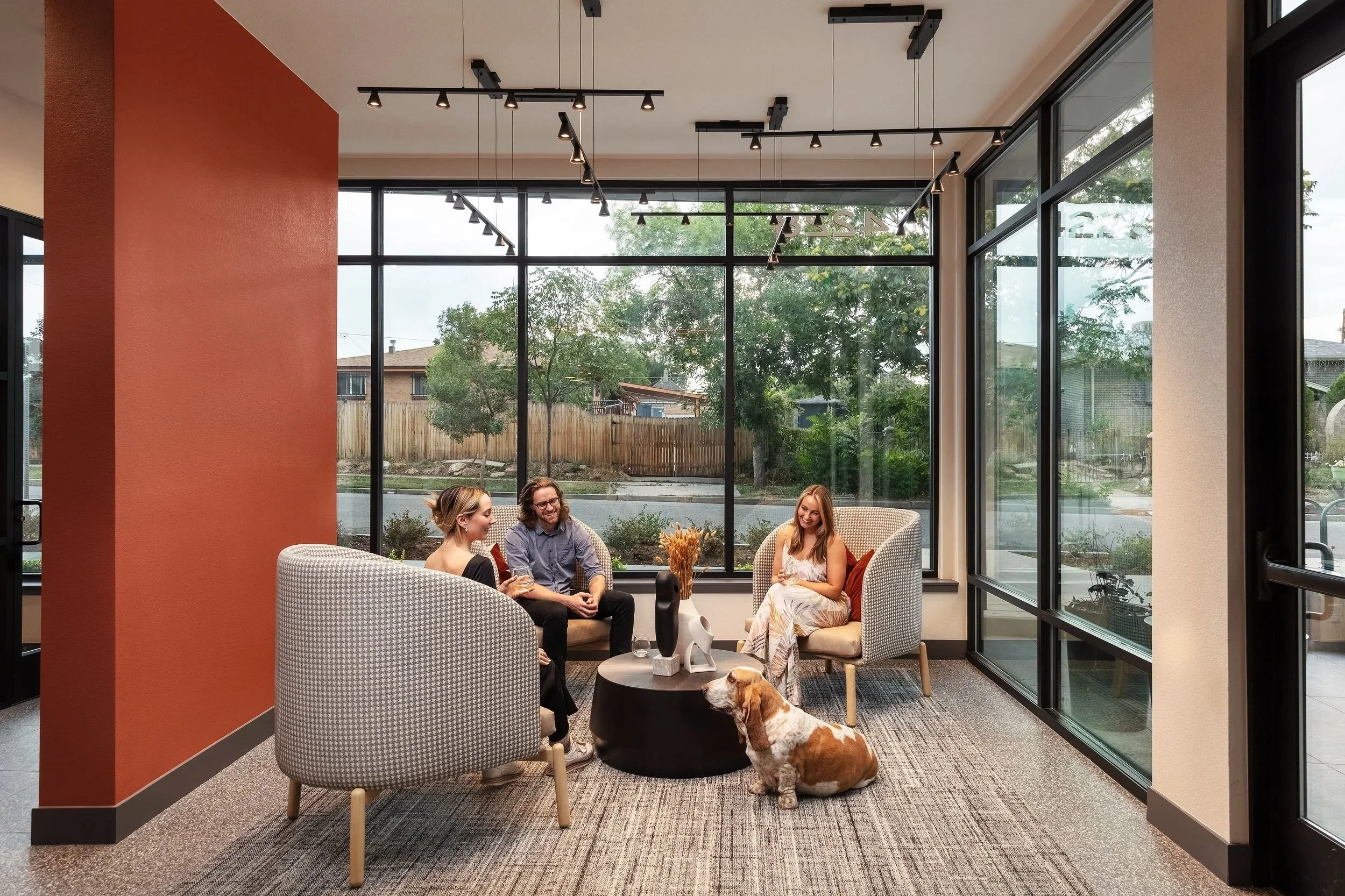 Four people sitting in a modern indoor lounge area with large windows, a small round black table with decorations, and a dog sitting on the carpeted floor.
